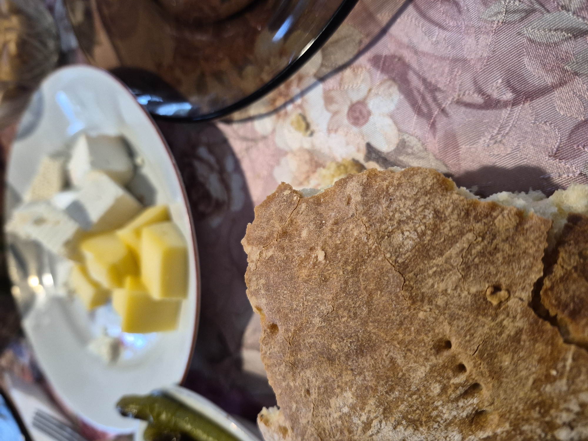 A close-up, slightly angled shot captures a table setting with food and a floral tablecloth. In the foreground, a piece of crusty bread with an uneven, textured surface is prominently displayed. The bread appears to be torn, revealing a soft, white interior. To the left of the bread, a white plate holds several cubes of cheese, some yellow and some white, arranged alongside what appear to be green pickled vegetables. A fork is partially visible to the upper left of the cheese plate. A dark-rimmed plate is visible on the right side of the image, reflecting light and obscuring its contents. The tablecloth beneath the food features a repeating floral pattern in shades of pink and purple. The scene suggests a meal is being served or has recently been enjoyed.