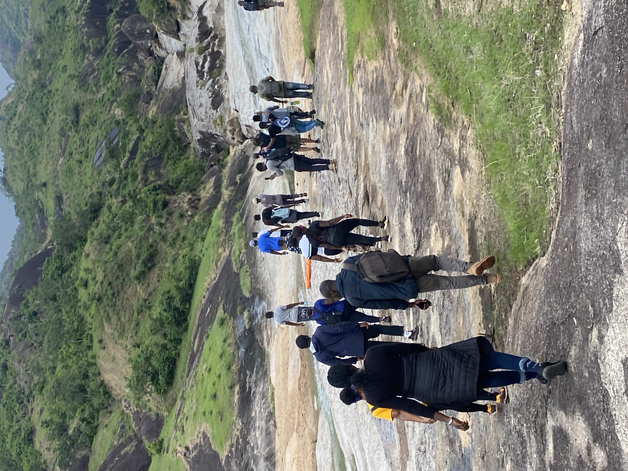 A group of approximately 18 people are standing or walking in a line on a large, rocky area. The rock is light colored and appears to be relatively smooth. To the left of the frame is a steep hillside covered in green vegetation. The sky is overcast. The people are wearing a variety of clothing. The scene appears to be in a natural, outdoor setting, most likely a rock outcropping or a mountainside location. It is in Ganawuri, Nigeria.