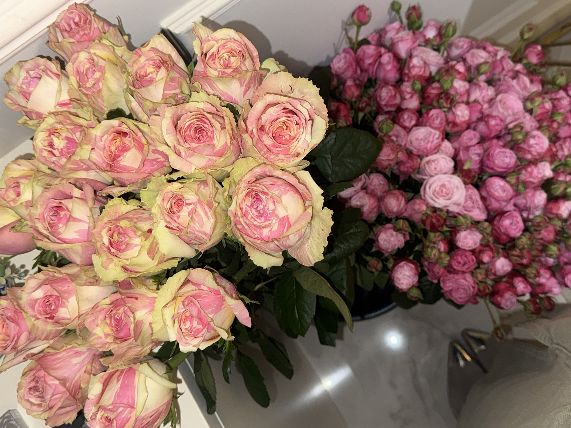 The image depicts two bouquets of flowers. One bouquet features large, pink and yellow variegated roses. The other bouquet consists of numerous small, pink roses. They are displayed indoors, likely on a table or countertop with a glossy surface reflecting light. The location cannot be determined to be Yerevan, Armenia, based solely on the image.
