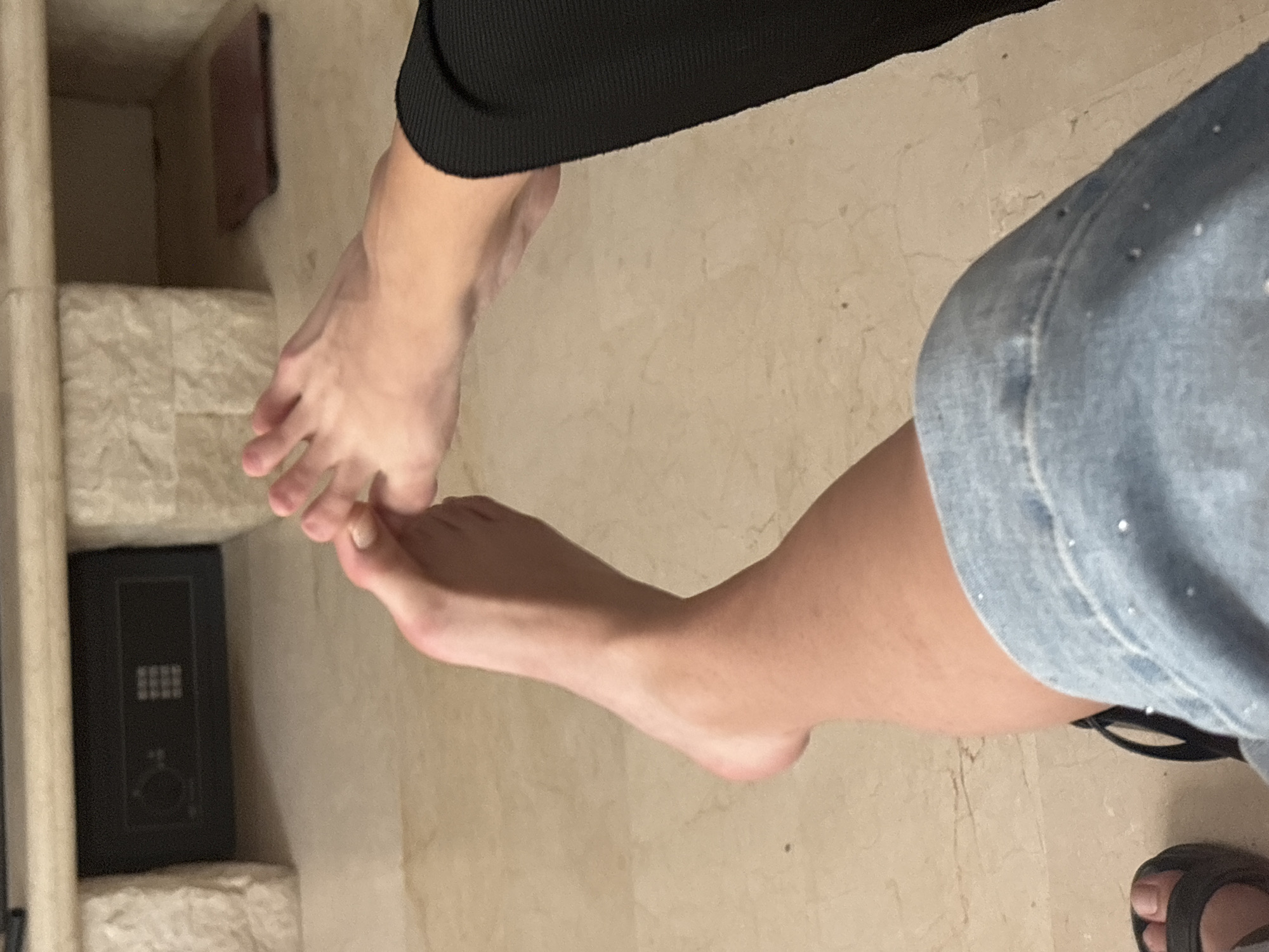 The image shows a close-up view of a person's feet. One foot is extended forward, and the toes are being touched by the fingers of the other hand, which is also visible in the frame. The person is wearing a denim skirt with some decorative elements. Part of a black sandal is visible on the left side of the image. The background appears to be an indoor setting with light-colored flooring and a stone-like structure. A black safe is embedded in the stone structure.