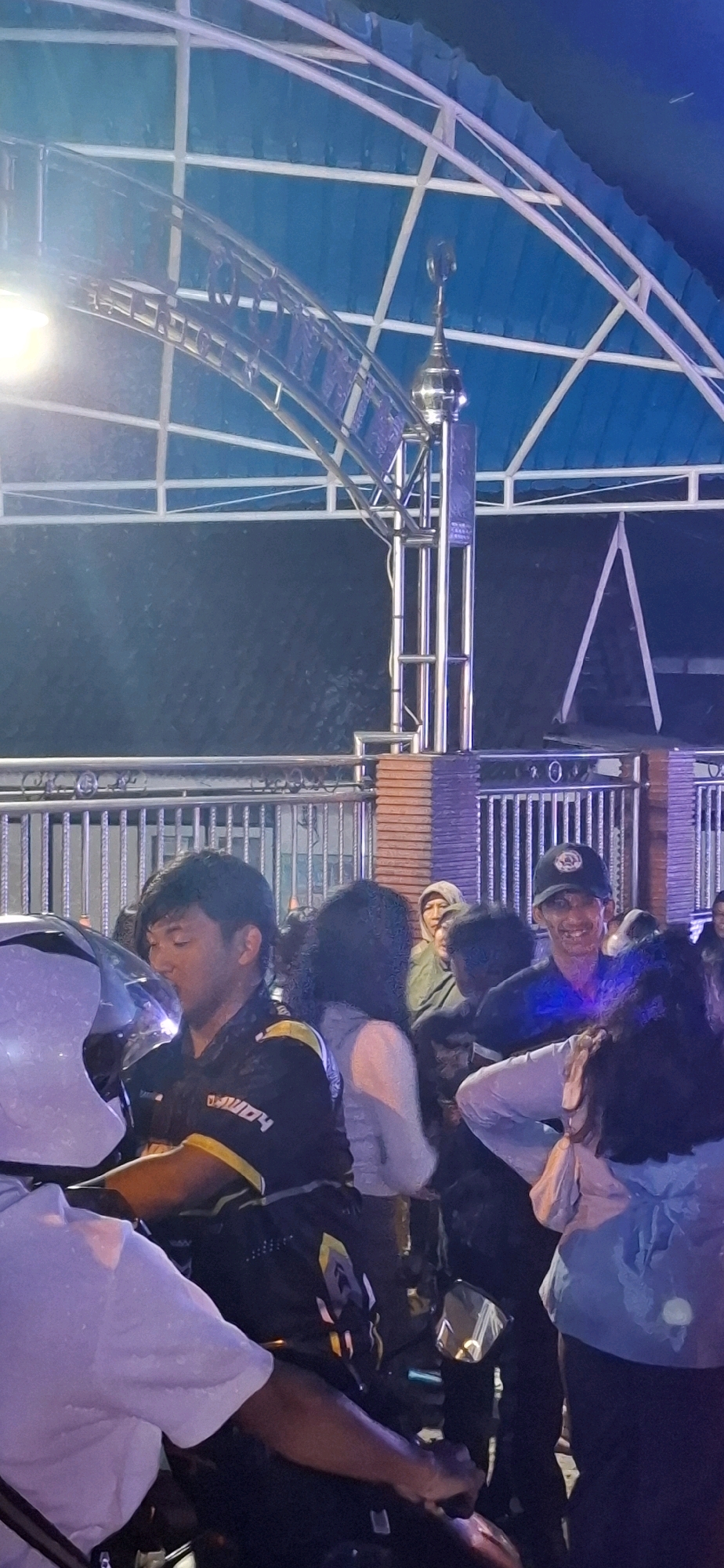 In Prigen, Indonesia, a group of individuals is gathered outdoors under a blue translucent roof structure, possibly a shelter or pavilion. The scene appears to be at night, with artificial lighting illuminating the area.
In the foreground, a person wearing a white t-shirt is partially visible on the left, with their arm resting on what seems to be a motorcycle. Next to them, a man in a black and yellow racing-style jacket is seated, with his head turned to the left. His jacket has text and logos on it, suggesting a connection to motorsports.
Behind these individuals, a crowd of people is visible. A man in a dark shirt and a black baseball cap is smiling and looking towards the viewer. Another person in a light-colored hooded garment, possibly a rain jacket, is looking forward. Other individuals are partially obscured by the foreground subjects and the crowd.
The environment features a metal fence and a brick wall in the background. The roof structure is supported by metal beams and appears to be a relatively modern construction. The overall impression is a gathering of people in a public or semi-public space, potentially during an event or casual meet-up.