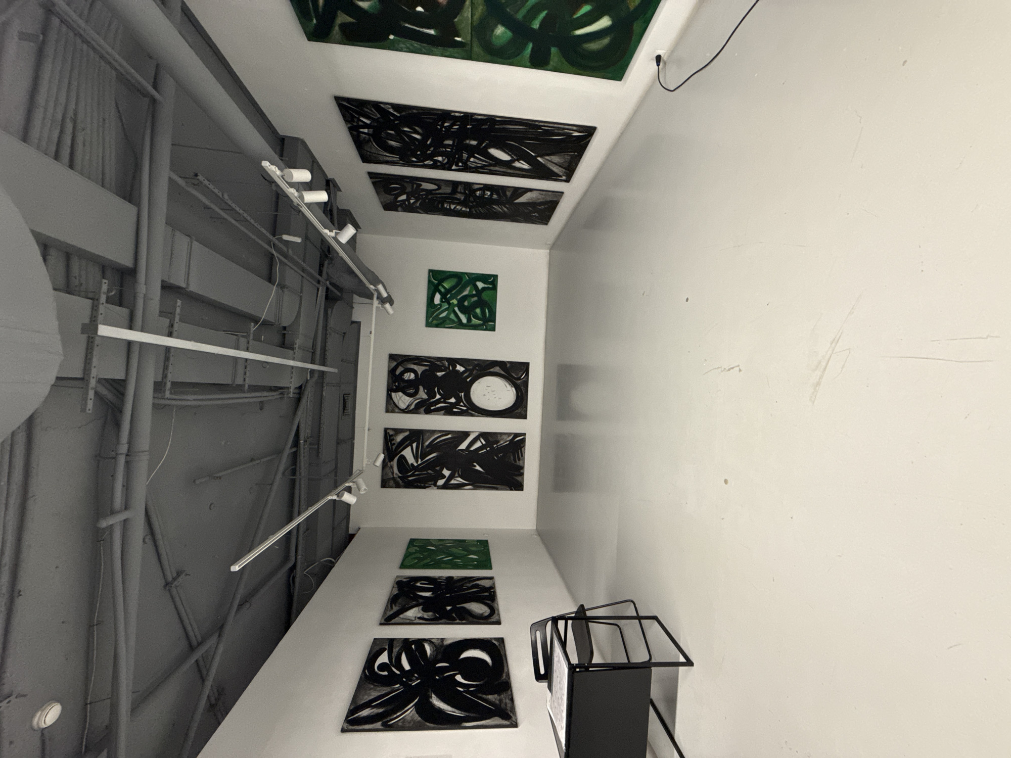 The image depicts an art gallery interior with a focus on the artworks displayed on the walls and ceiling. The space has a utilitarian feel, with exposed ceiling pipes and lighting fixtures. The art consists of a mix of abstract paintings with dark and green colors, and is displayed both on the walls and ceiling. There is a black metal stand in the lower right corner of the image. Without additional information, it is impossible to confirm if the scene is located in Moscow, Russia.