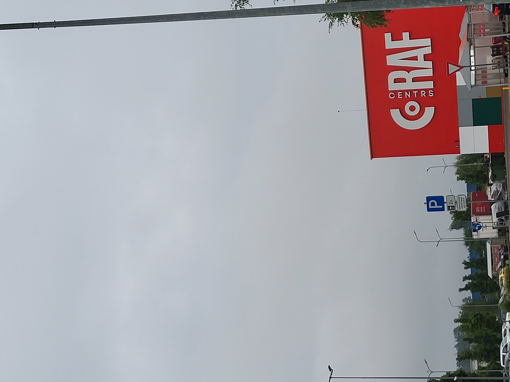 The image shows the RIO shopping center in Jelgava, Latvia. The shopping center's facade is painted bright red with the "RIO CENTRS" logo in large white letters. The sky is overcast. In the foreground, there are several parking lot features, including a traffic sign with a parking symbol and a "2h" time limit, streetlights, and a few parked cars. The scene depicts an urban environment with commercial buildings and infrastructure.