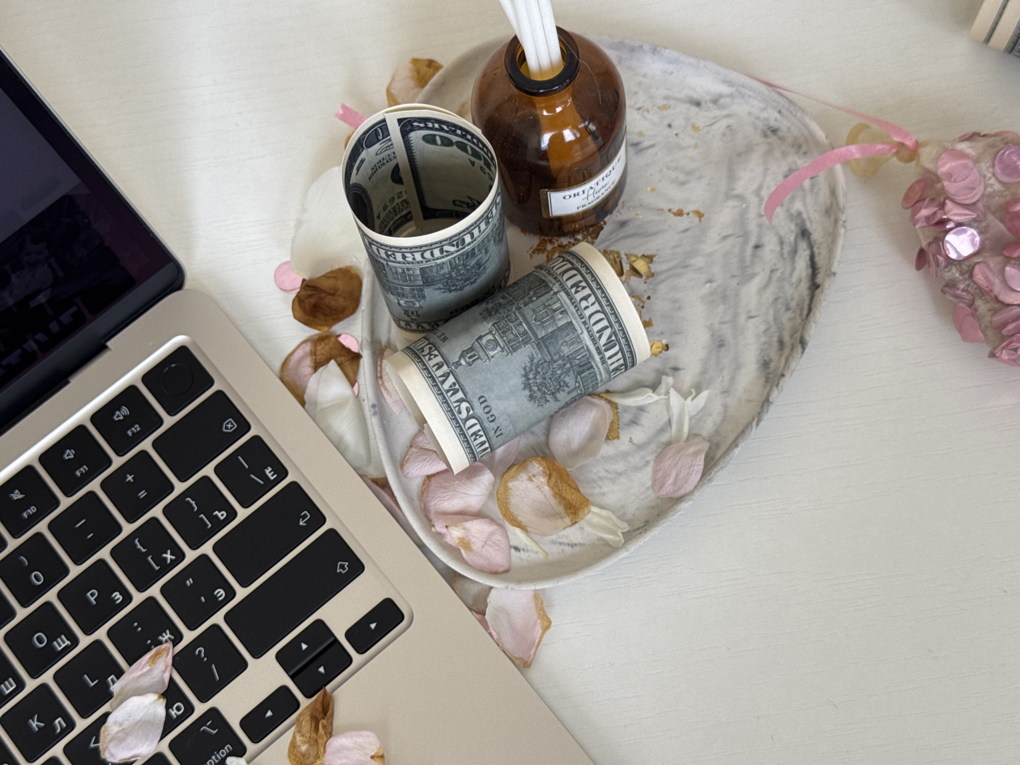 An overhead shot shows a laptop keyboard with scattered flower petals. Next to the laptop is a heart-shaped dish containing two rolled-up US dollar bills and a small amber-colored glass bottle with white reeds sticking out. The dish is also decorated with flower petals and gold flecks. The background is a plain white surface. The image does not contain any information or context that would allow for the determination of its location in the city of Yekaterinburg, Russia.