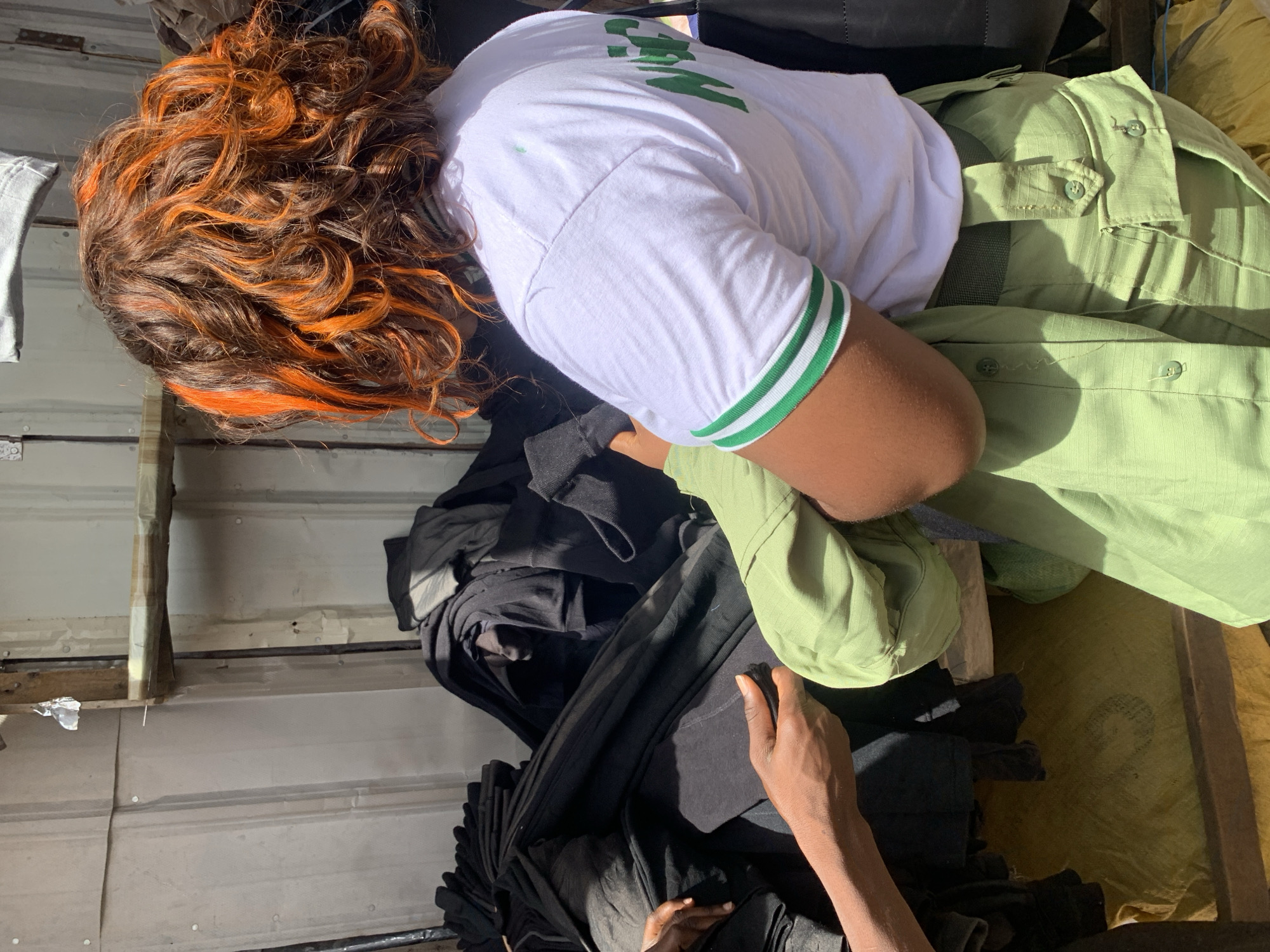 The image shows a person with dark brown hair with orange highlights lying down on a pile of clothes. The person is wearing a white t-shirt with green stripes on the sleeves and green lettering on the front. They are also wearing a green button-down shirt and matching green pants. The clothes are piled on a wooden pallet, which is placed on a yellow tarp. Some black clothes are piled around the person. The background is a corrugated metal wall. The setting appears to be an industrial or storage area, possibly in the city of Jos, Nigeria, based on the person's clothing (likely a National Youth Service Corps uniform) and the environment. A hand reaches into the frame from the bottom right.