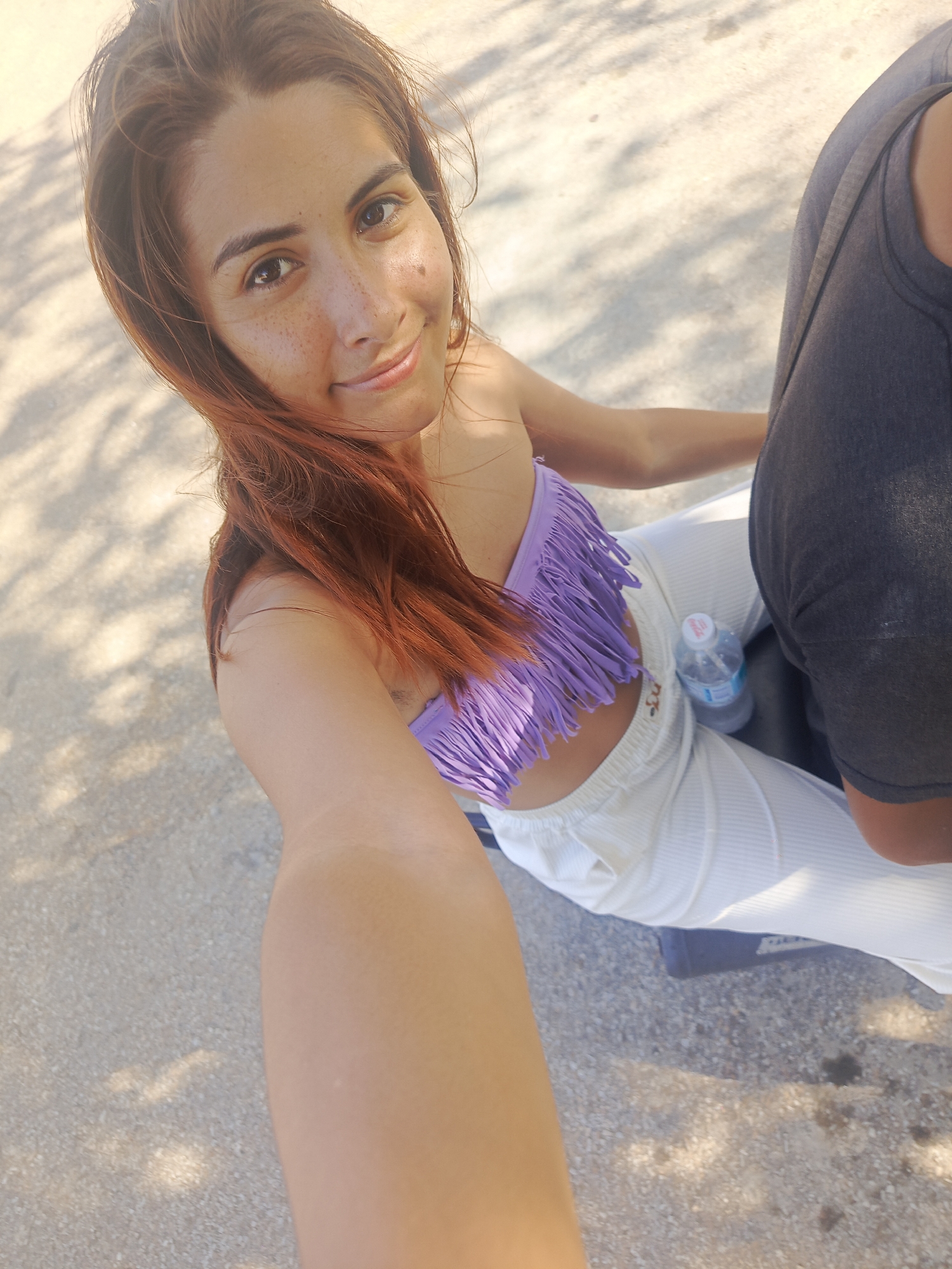 This is a selfie of a young woman with reddish-brown hair and fair skin, who has freckles across her nose and cheeks. She is smiling and looking at the camera. She is wearing a purple bikini top with fringe detailing and white, ribbed pants. Beside her is a person wearing a dark grey t-shirt. A bottle of water is visible on her lap. The scene appears to be outdoors, with a sandy or gravelly ground visible, and dappled sunlight suggesting it is daytime and possibly sunny. The shadows indicate foliage overhead. The context provided places the location in Pedro González, Venezuela. The overall mood is casual and relaxed.