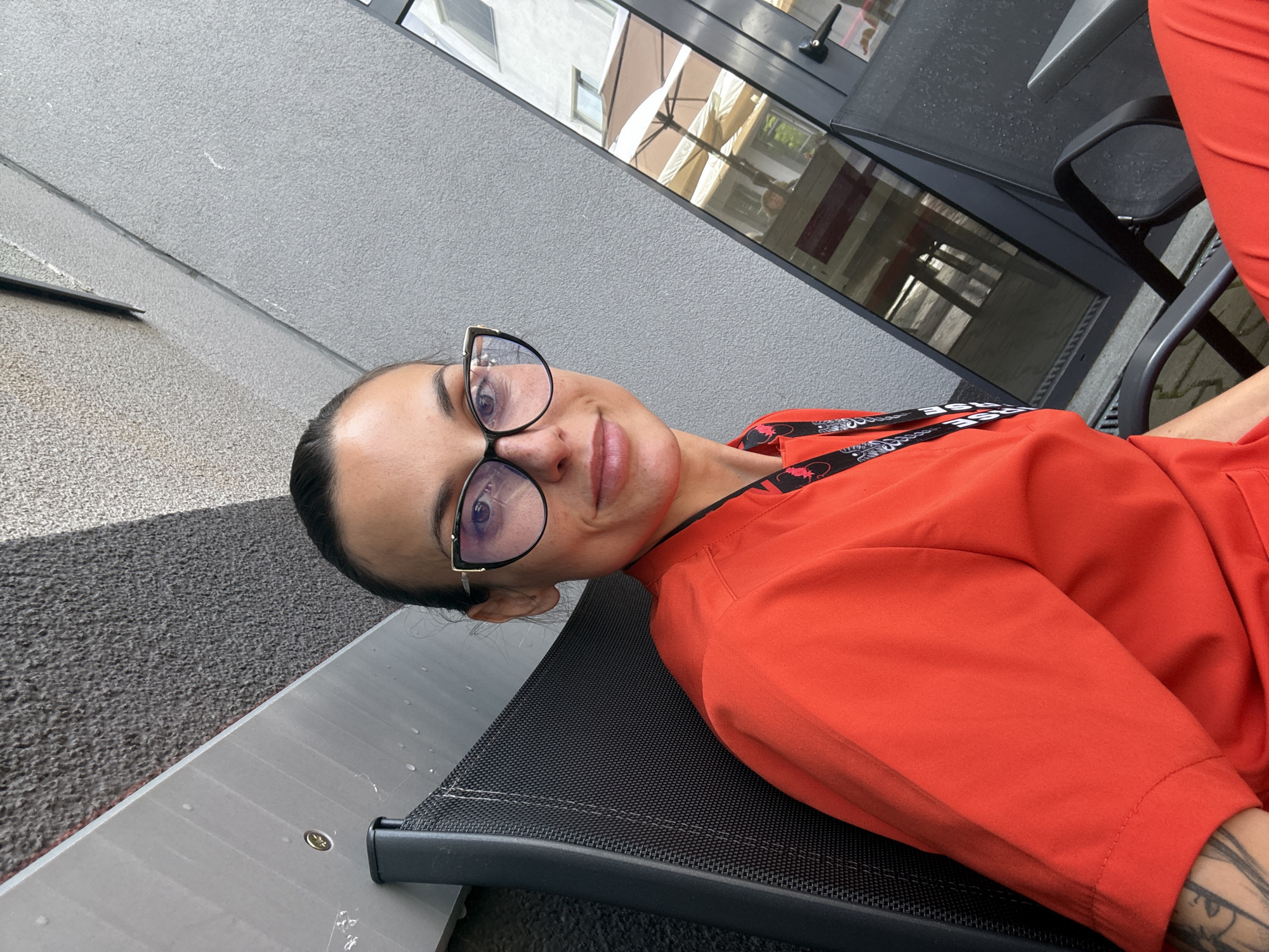 An eye-level, tilted outdoor shot features a woman from Pleven, Bulgaria, looking directly at the camera with a slight smile. She has dark hair pulled back and wears black and gold-framed glasses. Her attire includes a bright orange-red top and a black lanyard around her neck, which has "BASE" text and a red, patterned design, possibly featuring stylized dragons or similar motifs. Intricate tattoos are visible on her left arm.
The woman is seated on a black, mesh-like chair or bench. The background consists of a textured grey wall or ground surface, with a large window reflecting parts of a building, other windows, and possibly outdoor umbrellas, hinting at an urban or commercial environment. The lighting suggests it is daytime with soft, ambient light. Her expression indicates a relaxed and pleasant mood.