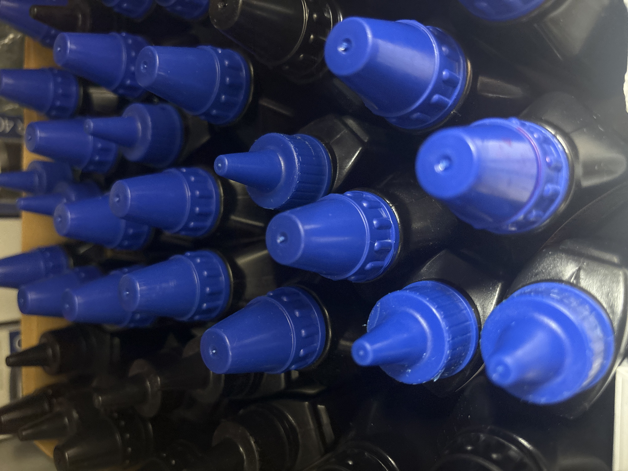 The image shows a close-up of numerous black bottles with blue caps. The bottles are densely packed together, and the perspective is slightly angled from above. The caps have a distinctive shape with a ridged design near the base and a conical top with a small hole. The bottles appear to be stored in a container or tray. Based on the prompt, the location is Chaikovsky, Russia. No people, actions, or interactions are visible in the image.