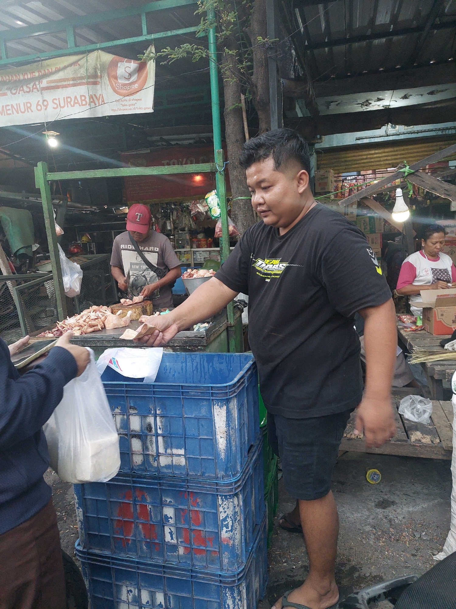 The image depicts an outdoor market scene, likely in Surabaya, Indonesia, with a focus on a food stall. In the foreground, a man in a black t-shirt and black shorts is holding a piece of paper and interacting with someone off-camera. Stacked blue plastic crates are visible to his left. Behind him, a butcher wearing a grey t-shirt and a red cap is cutting chicken on a wooden block, with a large pile of raw chicken in front of him. To the right, a woman in a pink and white shirt is also engaged in an activity at another stall. The market appears to be covered by a corrugated metal roof, with natural light filtering through. A banner advertising "SHOPEE" and "MENUR 69 SURABAYA" is prominently displayed in the background. The ground is paved with rough concrete.