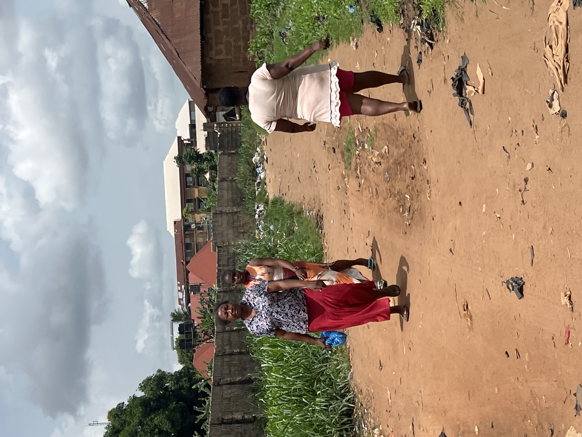 Three women walk along a dirt road lined with garbage and lush vegetation in Obosi, Nigeria. The first woman is wearing a blue floral shirt, red pants, and sandals and is carrying a blue bag. The second woman behind her is wearing an orange dress and blue sandals. The third woman, further up the road, is wearing a beige shirt and red shorts. A concrete wall runs along the left side of the road with buildings visible behind it. The sky is overcast.
