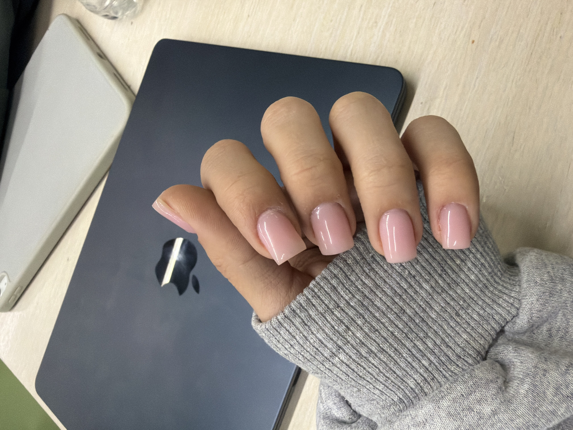 A human hand, adorned with short, square-shaped fingernails coated in a semi-opaque pink polish, rests on a dark blue Apple iPad. The hand is partially covered by the cuff of a grey knit sweater. The iPad sits next to a white electronic device on a light-colored, possibly wooden, surface. No specific location details or indications of Yerevan, Armenia, are discernible from the image.