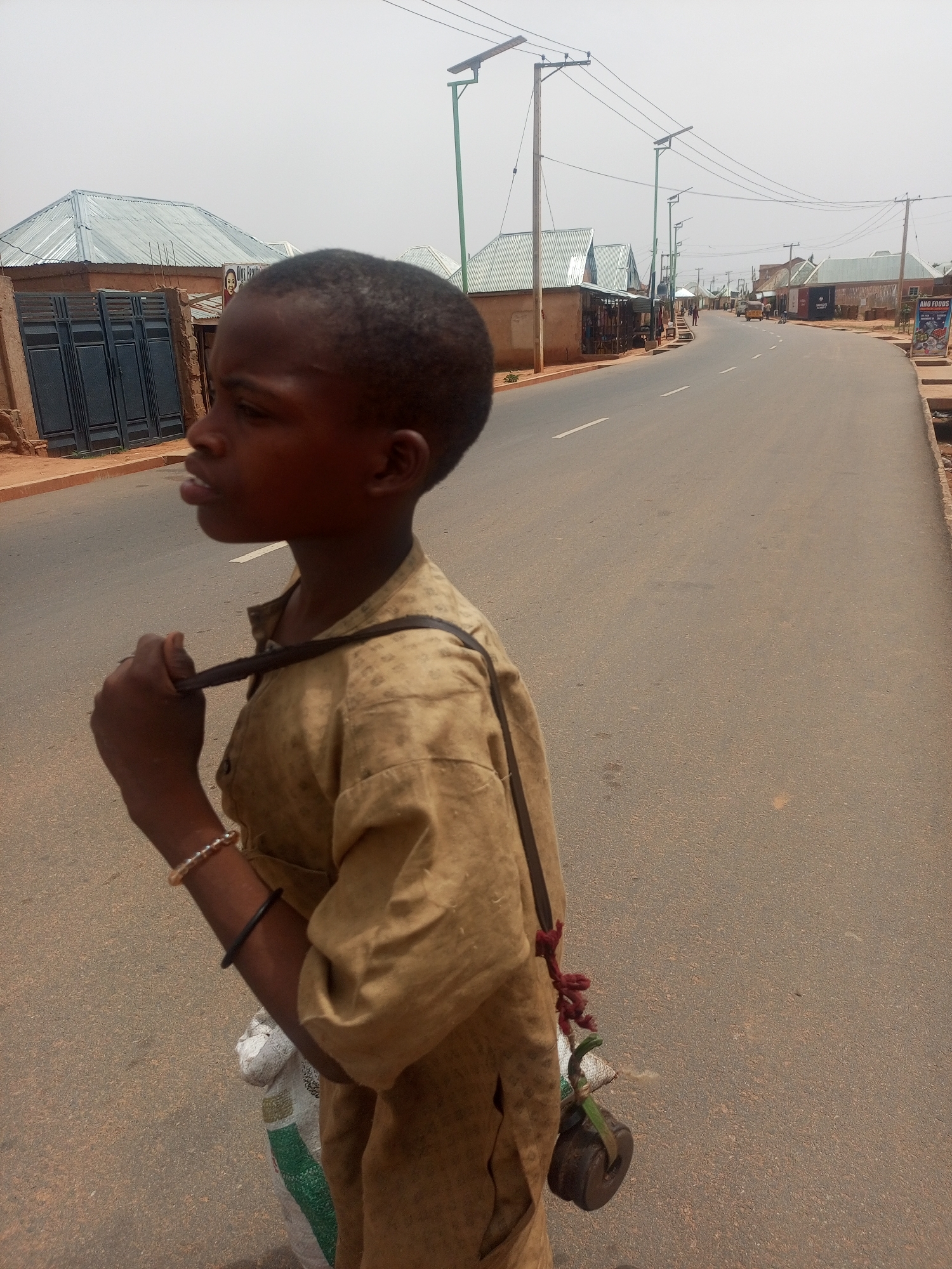 A young boy stands on the edge of a paved road in Tempure, Nigeria. He is wearing a tan shirt and has a bag slung over his shoulder. The road stretches into the distance with buildings on either side. Utility poles line the street.