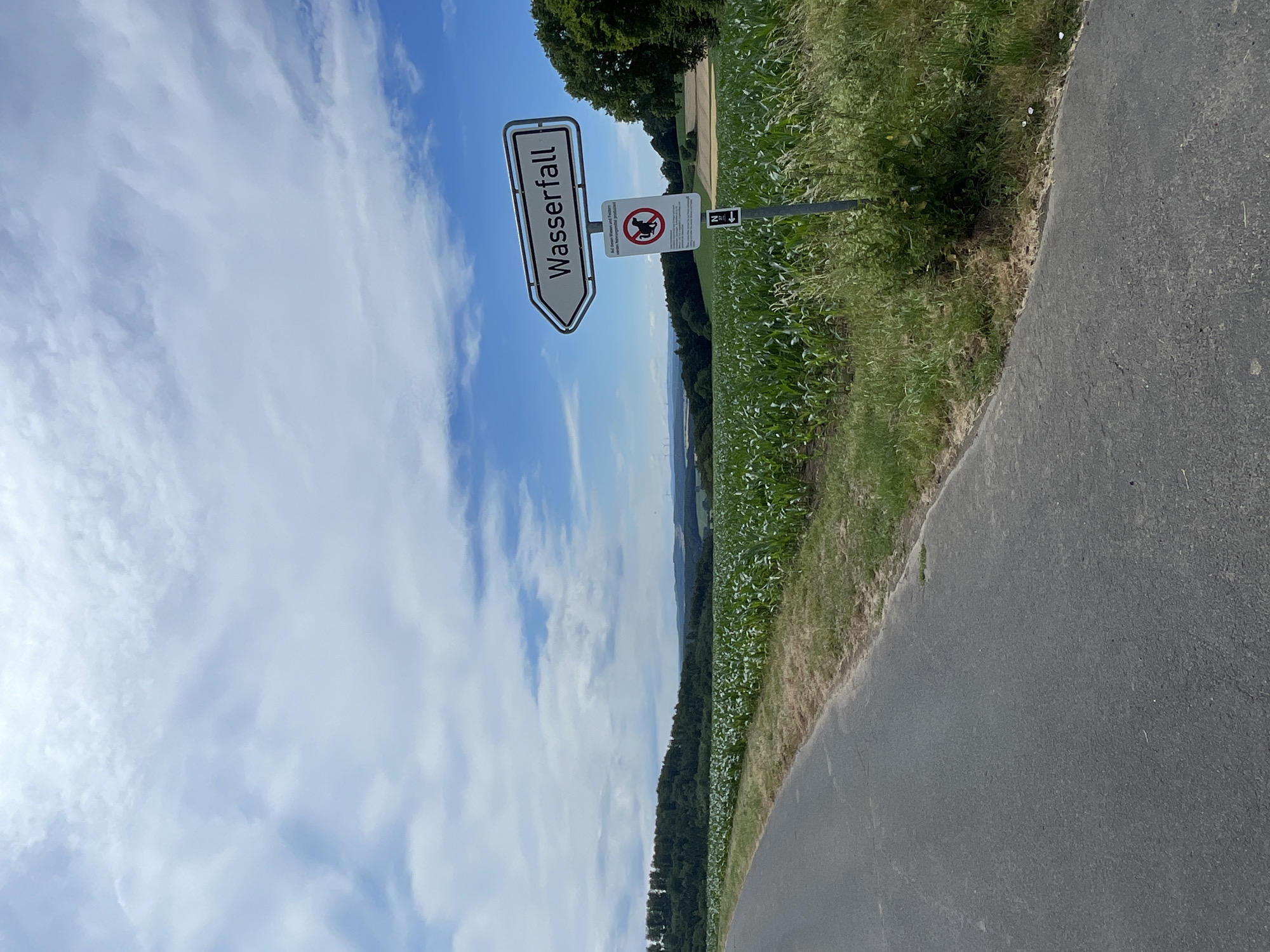 This is a landscape photograph taken in Christerode, Germany. The image features a road curving to the right, bordered by lush green grass and a field of crops. In the background, a dense forest stretches across rolling hills under a bright blue sky dotted with fluffy white clouds.
Dominating the left side of the frame is a white directional sign with a black border pointing to the left. The German word "Wasserfall" (waterfall) is written in black text on the sign. Next to it, on a separate pole, is a circular sign with a red border and a diagonal line across it, indicating prohibition. Inside the circle, a black silhouette of a dog is depicted, with text below it reading "Auf privaten Wiesen und Feldern..." which translates to "On private meadows and fields...". Another smaller sign with a black arrow pointing downwards is also visible on the same pole.
The scene suggests a pleasant daytime outing, possibly for hiking or cycling, given the road and directional signage. The weather appears to be clear and sunny, conducive to outdoor activities. The time of day is likely midday or early afternoon, based on the bright sunlight. The overall mood is one of tranquility and natural beauty, inviting exploration of the surrounding landscape.