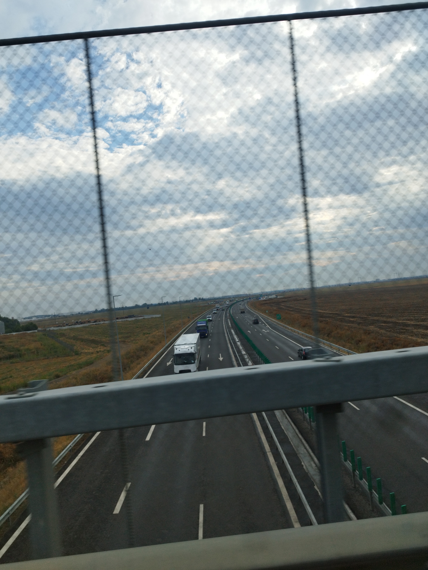 The image shows a view of a highway in Popești-Leordeni, Romania, from an elevated position, likely a bridge, looking through a wire fence. The highway has multiple lanes and is busy with traffic, including trucks and cars. The surrounding landscape is flat with agricultural fields and some industrial structures visible in the distance. The sky is overcast with clouds. The foreground is dominated by the metal railing and wire fence, which are slightly out of focus, framing the highway scene.