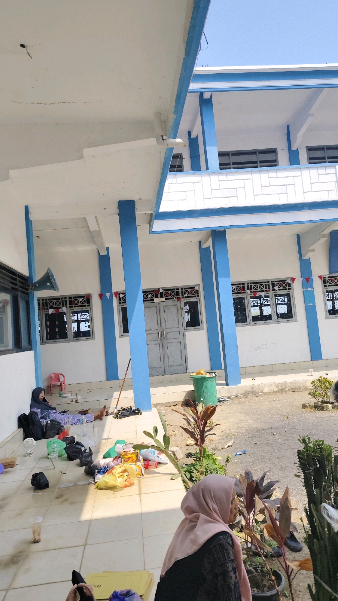 The image depicts an outdoor area in front of a two-story building with blue and white accents. The building appears to be a school or similar institutional structure, with multiple windows and doorways visible on the ground floor. In the foreground, several people are present. One individual, wearing a light pink hijab and a patterned dress, is seated with their back to the camera, looking towards the right. Another person, also wearing a hijab and dark clothing, is lying down on the tiled ground on the left side of the image, near a collection of bags and items. A red plastic chair is visible nearby. The ground is tiled in the immediate vicinity of the building, transitioning to gravel and dirt further away. Various objects are scattered on the ground, including black trash bags, grocery bags, plastic cups, and a green trash can. Red, white, and blue triangular flags are strung along the front of the building above the windows, and some are also attached to the window frames. The sky is clear and blue. The overall impression is of a casual, possibly waiting or resting area outside a building. The presence of flags suggests a celebratory or national occasion.