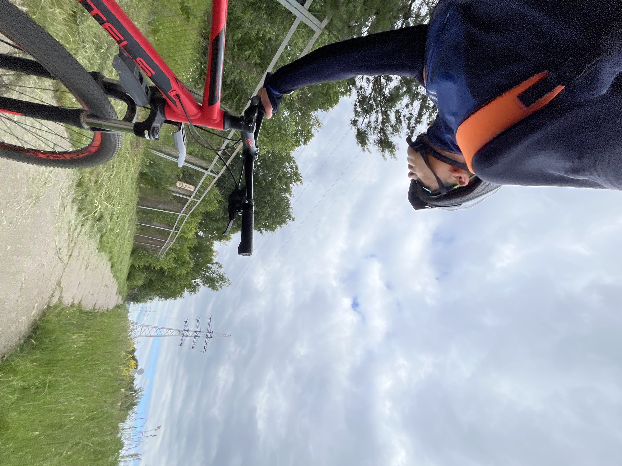 A first-person perspective shot shows a person in cycling attire and a helmet leaning forward, holding the handlebars of a red and black mountain bike. The bike's front fork and wheel are visible in the foreground. The path ahead is made of dirt and is flanked by tall green grass. In the background, a metal fence runs along the left side, with trees and foliage behind it. A tall, lattice-like power line tower stands in the distance on the right, under a cloudy sky with patches of blue. The scene appears to be an outdoor trail, possibly on the outskirts of Chaikovsky, Russia, given the presence of power lines and the natural landscape. The overall impression is of a moment captured during a cycling excursion.