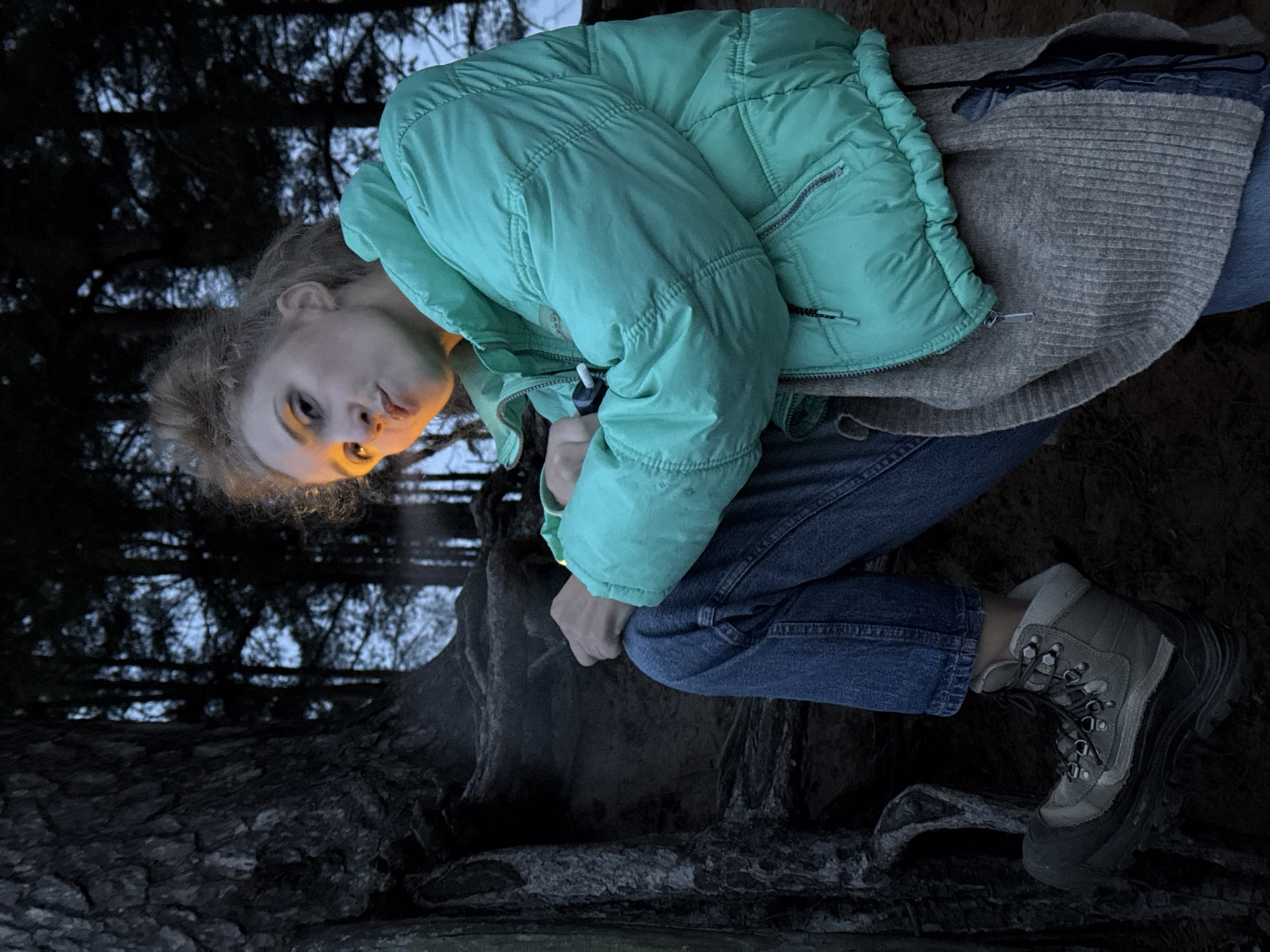 This is a low-light, portrait-oriented full shot depicting a person in a forest environment, likely in Chaikovsky, Russia. The image appears to be taken during dusk or night, with the natural light fading.
The subject is a young person with light-colored hair, wearing a teal or mint green puffer jacket over a grey, ribbed knitted cardigan, blue jeans, and sturdy light grey and black hiking boots. They are in a crouched or seated position on the dark ground, which is covered with visible tree roots and natural debris. A small, white cylindrical object is held in their right hand.
The most prominent feature is a strong, warm light source originating from below and to the side of their face, illuminating their features dramatically and casting distinct shadows. This artificial light highlights their expression, which appears intense and contemplative as they look directly at the viewer.
The background is dominated by the dark, silhouetted forms of tree trunks and branches, with a pale, twilight sky visible through the sparse foliage above. The person's attire suggests cool or cold weather conditions, suitable for an outdoor setting in a wooded area.