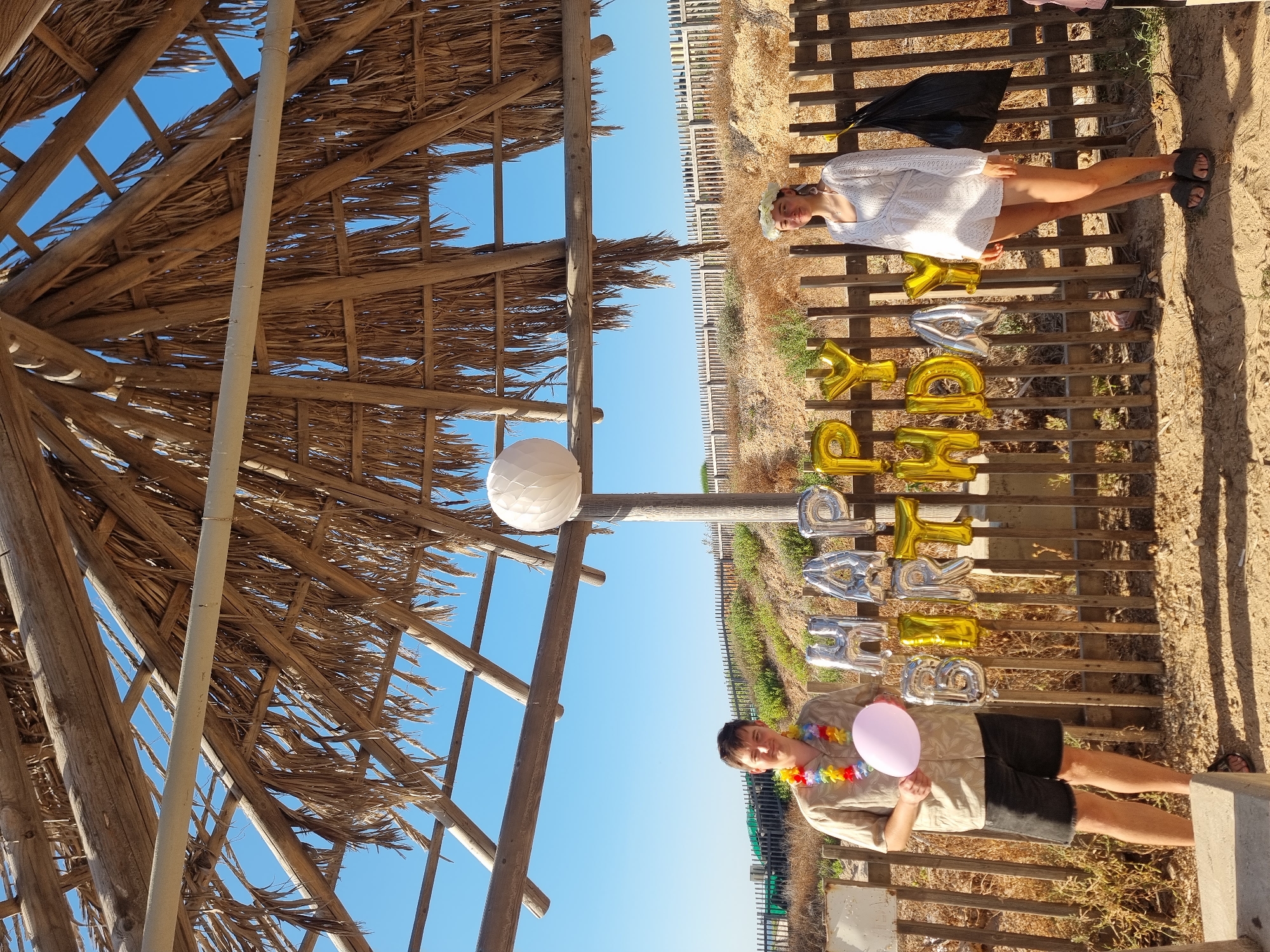 In Palmachim, Israel, two people pose for a photograph under a thatched-roof shelter, likely on a beach or near one. A man, wearing a Hawaiian shirt, lei, and dark shorts, stands on the left, holding a pink balloon. To his right, a woman in a white dress and flower crown stands. Behind them, a wooden fence displays "HAPPY BIRTHDAY" balloons, a mix of silver and gold. The environment is dry and sandy with sparse vegetation, indicating a coastal or arid climate. A clear blue sky dominates the background.