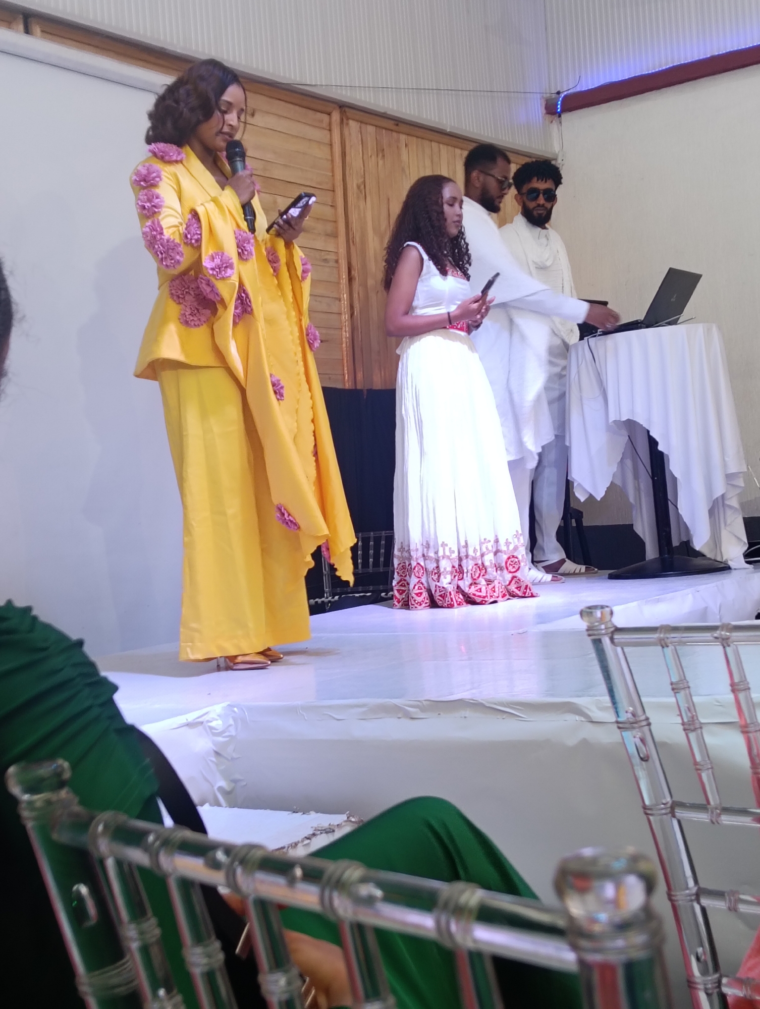 A full shot depicts three individuals and part of an audience on a stage in a location in the city of Unknown, Unknown. The foreground is filled with a portion of the audience, including a person in a green dress and a metal and clear plastic chair. The stage is covered in white fabric.
A woman in a vibrant yellow suit with pink floral appliqués on the shoulders and arms stands on the left side of the stage, holding a microphone in her left hand and a mobile phone in her right. Beside her is a woman in a traditional white Ethiopian dress with red geometric patterns at the bottom, holding a mobile phone in her left hand. Next to her, a man in a white ensemble with sunglasses is working on a laptop placed on a small table draped with a white cloth. The man in the middle is dressed in a white suit. The backdrop consists of a partially visible wooden panel. Overhead lighting is present, emitting a soft glow on the scene.