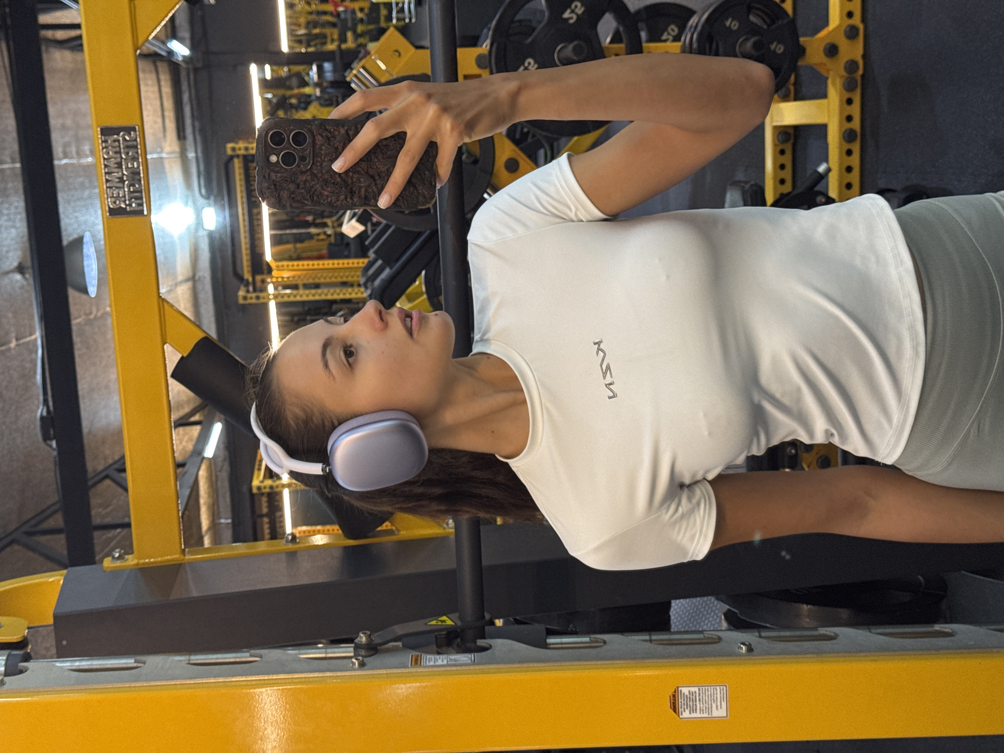 A young woman is taking a selfie in an indoor gym in Chalong, Thailand. She is positioned next to bright yellow and black gym equipment, holding a dark, textured smartphone with triple camera lenses in her right hand, pointed towards herself. She wears light purple over-ear headphones, a white short-sleeved athletic top with a reversed logo resembling "Ksen," and grey athletic leggings.
The gym environment features yellow and black exercise machines and racks. In the background, several weight plates are visible on a rack, marked with "50," "40," and "10." A metallic plaque on a yellow support beam displays the reversed text "COMMODORE FITNESS," while a "WARNING" label is partially visible on another yellow surface. The gym is brightly lit with artificial lighting. The woman has a focused expression, seemingly pausing during her workout to capture the moment.