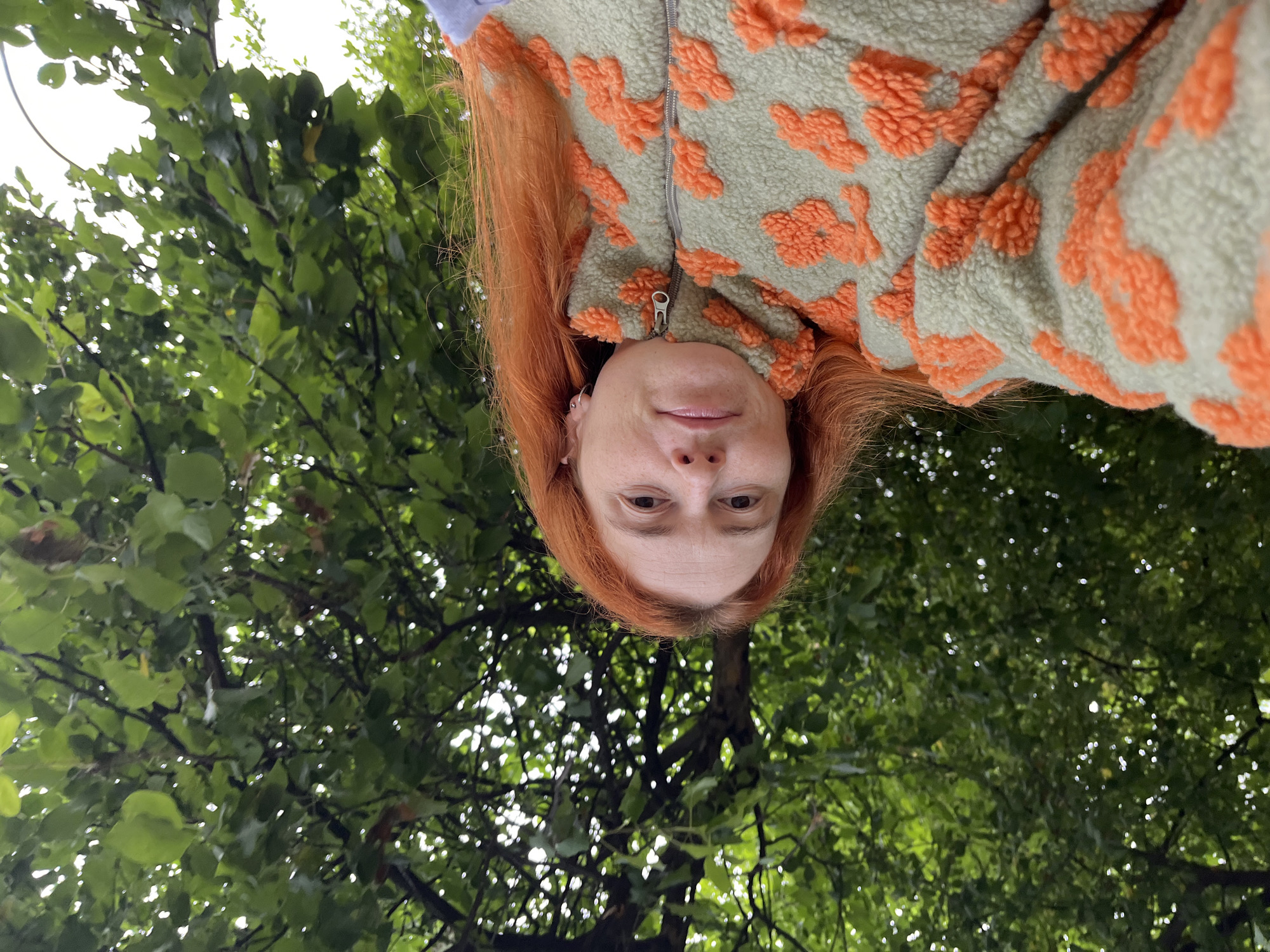 A woman with long, bright orange hair is captured in a close-up selfie taken outdoors. She is wearing a fuzzy light green jacket with an orange floral pattern and a zipper. The background is dominated by the lush green leaves and branches of a tree, suggesting a park or natural setting. The image does not provide any clues about the specific location within the city of Chaikovsky, Russia. The lighting is soft, indicating an overcast day or shade.