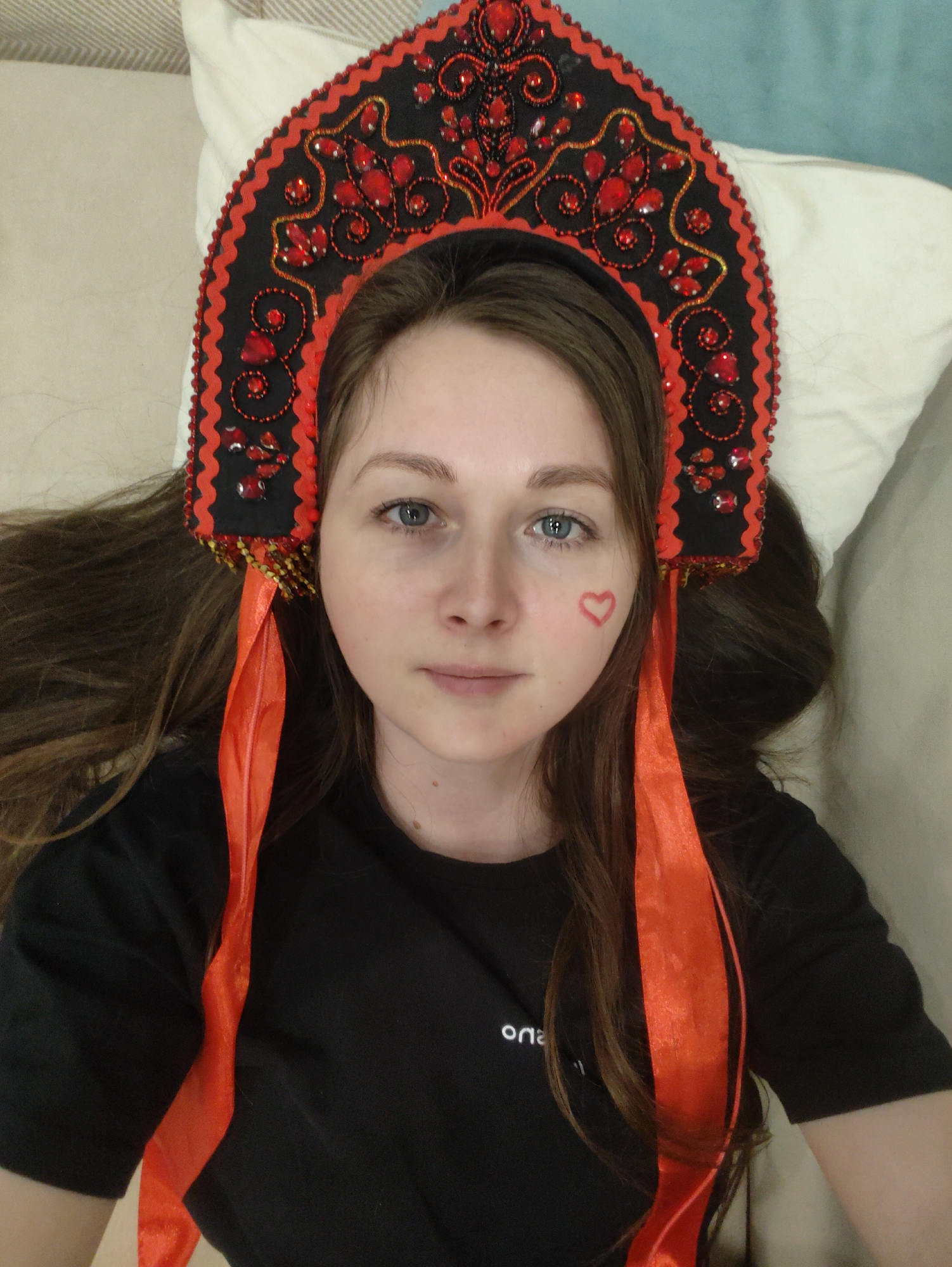 A woman lies on a light-colored surface. She has long brown hair, fair skin, and blue eyes. On her right cheek is a red heart drawn on her skin. She is wearing a black shirt with a small white logo or text on the left side. On her head, she wears a black headdress decorated with red beads, stones, and gold trim. Two wide, red ribbons extend from the sides of the headdress down along her shoulders. The background consists of a light-colored surface, possibly a sofa or bed. Location: Unknown, Unknown.