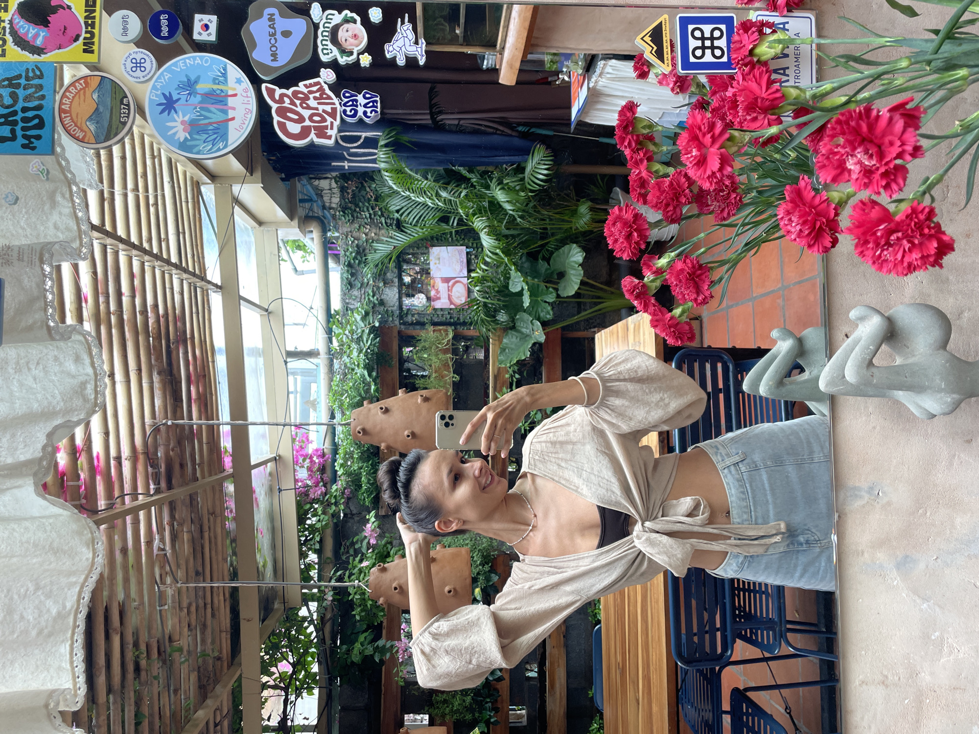 A woman with her hair in a bun, wearing a beige cropped blouse tied at the front and a light blue denim skirt, takes a mirror selfie. She is standing indoors, in what appears to be a cafe or restaurant in Mui Ne, Vietnam. The cafe has a rustic ceiling made of bamboo. In the background, there are tables, chairs, and lush green plants. A bouquet of bright pink carnations is prominently displayed in the foreground, reflected in the mirror along with the woman and parts of the cafe's interior. Various stickers are visible on the glass surface of the mirror, including one featuring a mountain range labeled "MOUNT ARARAT 5137 m" and another with a Korean flag.
