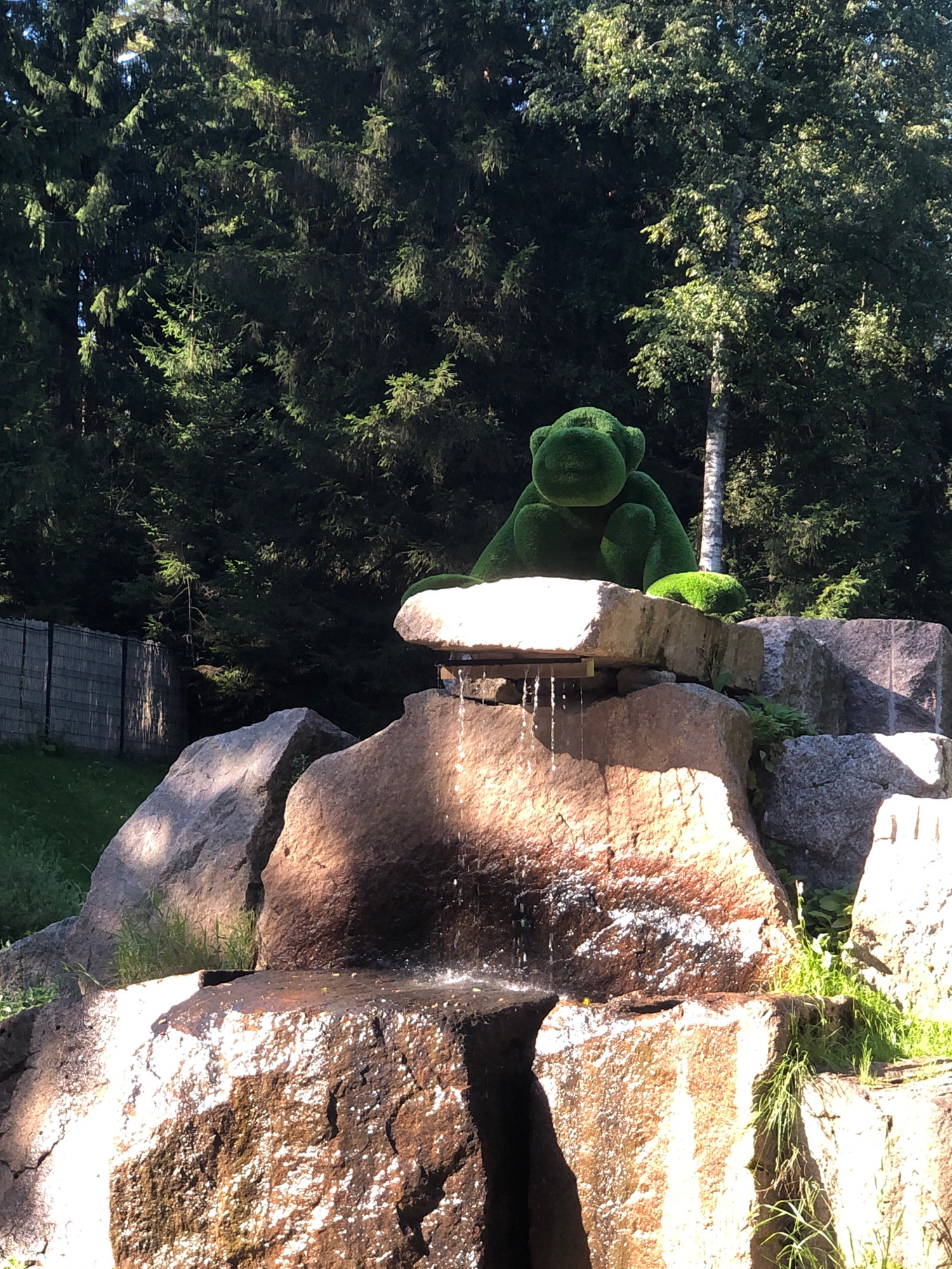 In Okhtinskiy Park, Russia, a green topiary sculpture resembling a monkey sits on a rock formation. The sculpture is positioned centrally, with the monkey appearing to be in a seated pose. Below the monkey, water cascades down the rocks, forming a small waterfall. The surrounding environment is lush and green, with a dense forest of trees forming the backdrop. The sunlight filters through the trees, casting dappled shadows on the rocks and sculptures. A fence is visible on the left side of the image, and grass covers the ground.