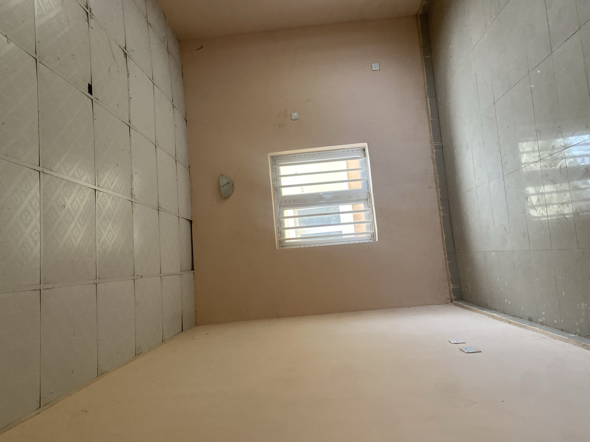 The image shows an interior view of an empty room in Bulamare, Nigeria. The room has light pink walls and a tiled floor in a neutral grey color. A white-framed window with vertical bars is visible on one wall, and it appears to be a double-pane window. On the same wall as the window, there are two electrical outlets. Another electrical outlet is visible on the opposite wall. The ceiling is made of square tiles, some of which appear to be damaged or discolored. A light fixture is attached to the ceiling near the window. The room appears to be recently painted or under renovation, as there are some drips of white paint along the base of the walls. There are no people or furniture visible in the room. The lighting suggests it is daytime, with natural light entering from the window.