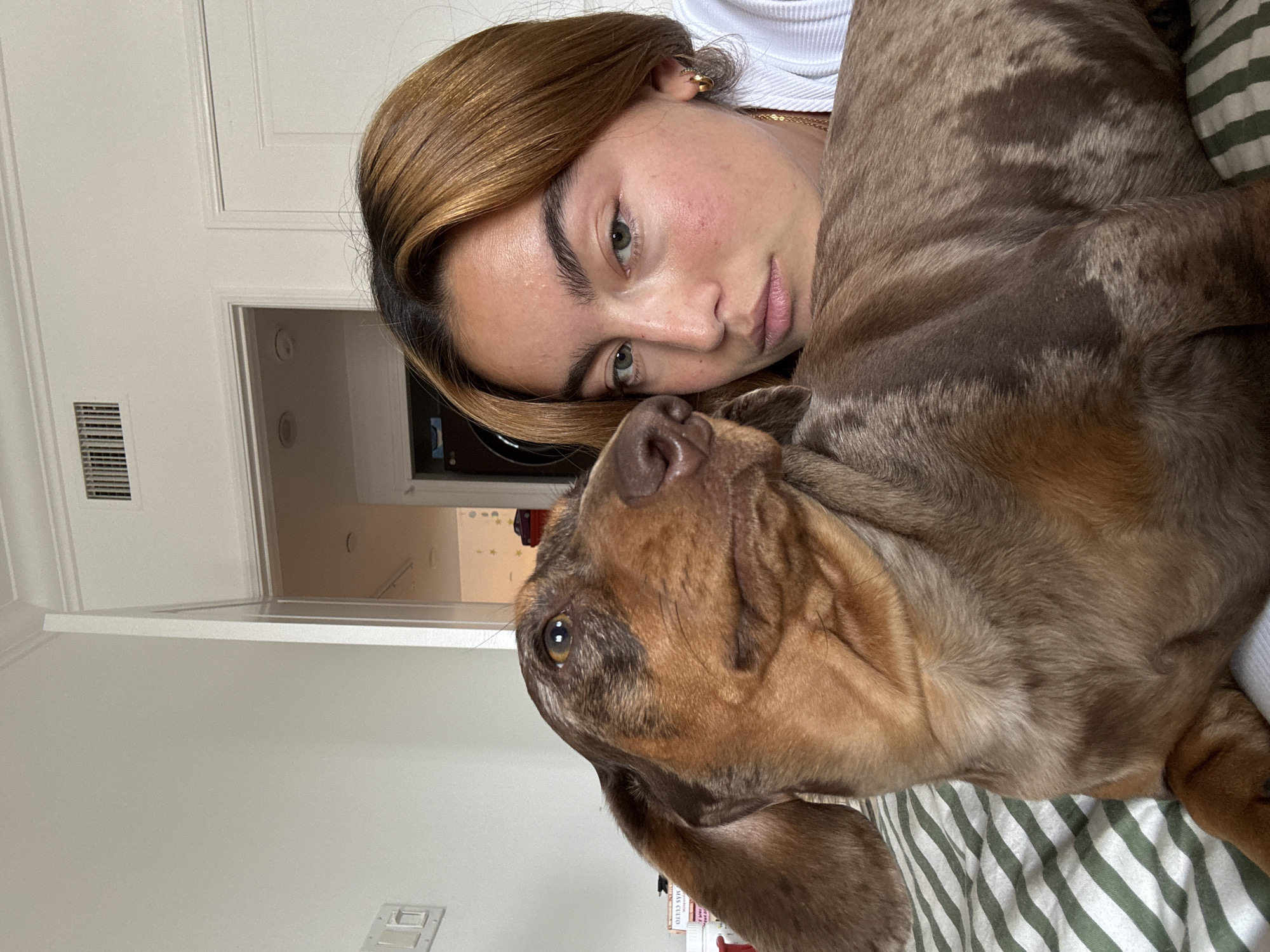 The image shows a young woman with light brown hair lying in bed with a brown and tan-colored dog. The woman's face is visible, and she appears to be looking directly at the camera. The dog is lying on top of her, with its head resting on her chest. The bed has a green and white striped comforter. The background shows a room with white walls and a partially visible doorway. The location is likely a residential interior in Los Angeles, United States of America.