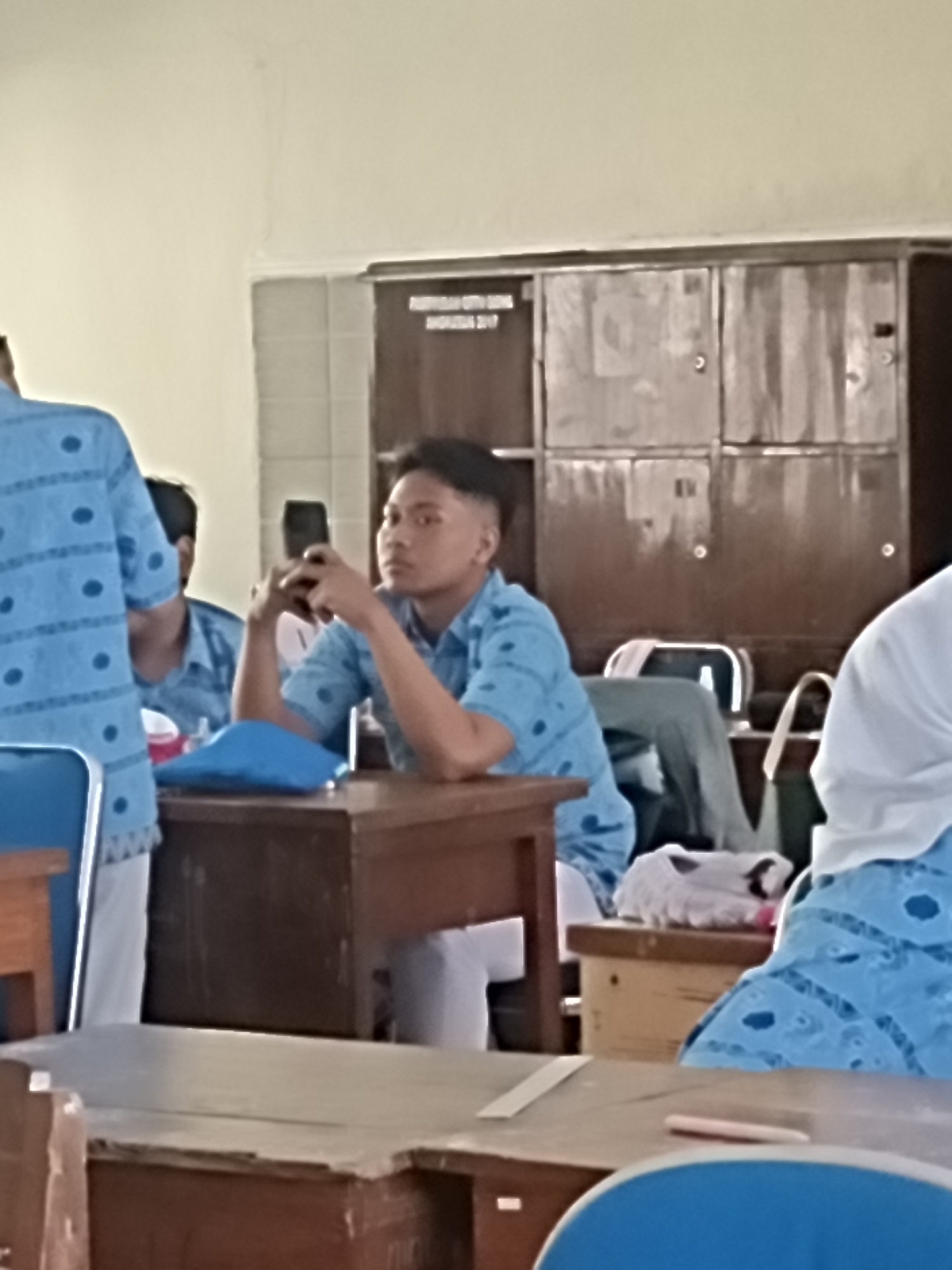The image depicts a classroom scene in Kediri City, Indonesia, with several students present. In the foreground, a young male student is seated at a wooden desk, holding a smartphone in his hands and looking towards it. He is wearing a blue patterned short-sleeved shirt, likely a school uniform. To his left, another student, partially visible, is also wearing a similar blue patterned shirt. A portion of a third student, wearing a white head covering and a blue patterned garment, is visible on the right side of the image. Behind the seated student, a set of lockers or cabinets are mounted on the wall. The environment appears to be a classroom, suggested by the desks, chairs, and lockers, common in Indonesian educational institutions. The lighting suggests it is daytime, possibly indoors with artificial or natural light.