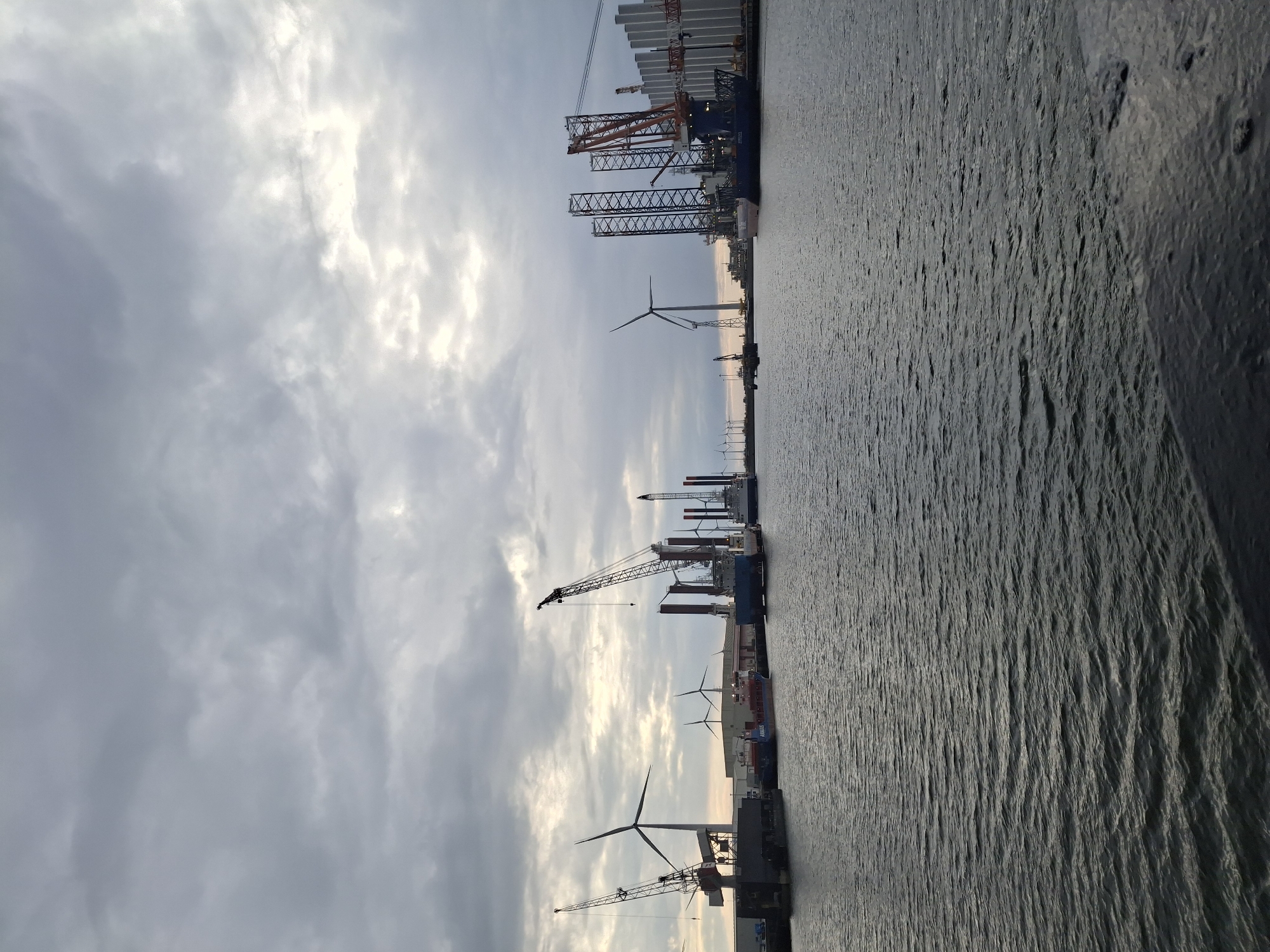 The image depicts a waterfront scene in Eemshaven, Netherlands, with a cloudy sky overhead. Several large cranes and wind turbines are visible along the harbor, suggesting an industrial area focused on renewable energy infrastructure. The water appears calm, reflecting the overcast sky. The foreground features a concrete or stone structure from which the photograph was likely taken. There are no people visible in the frame.