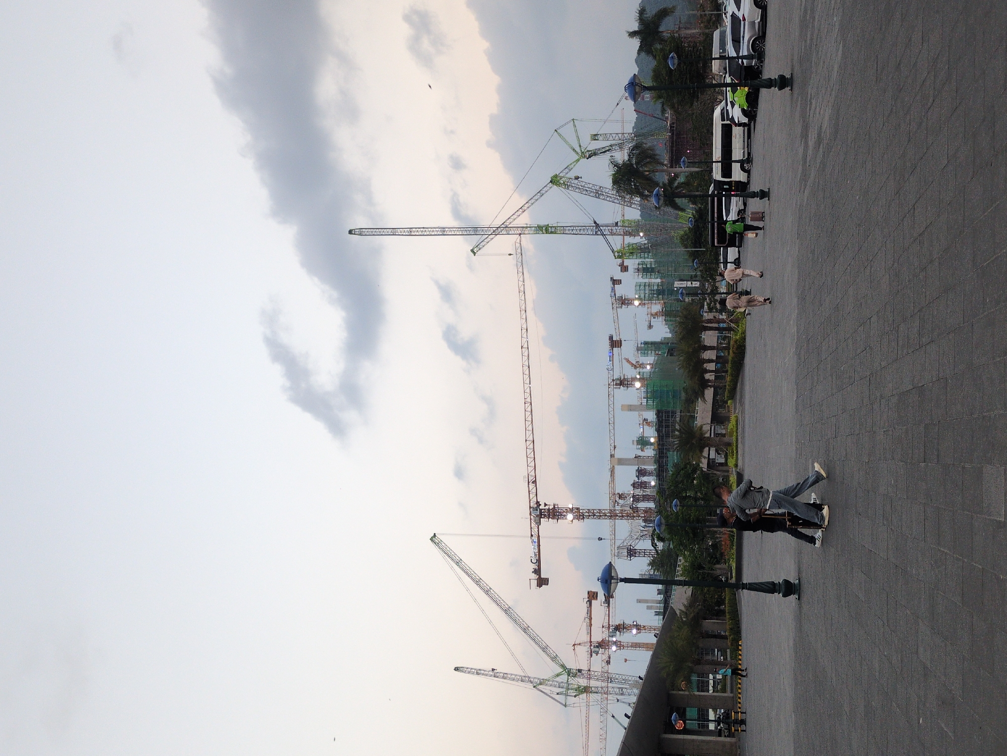 The scene is in Phú Quốc, Vietnam. The environment is an urban area featuring numerous construction cranes against a cloudy sky. In the foreground, there is a paved area with street lamps. Several people are present, including a couple, who are embracing. Additionally, there are pedestrians walking in the distance. Several cars and vans are also parked in the background.