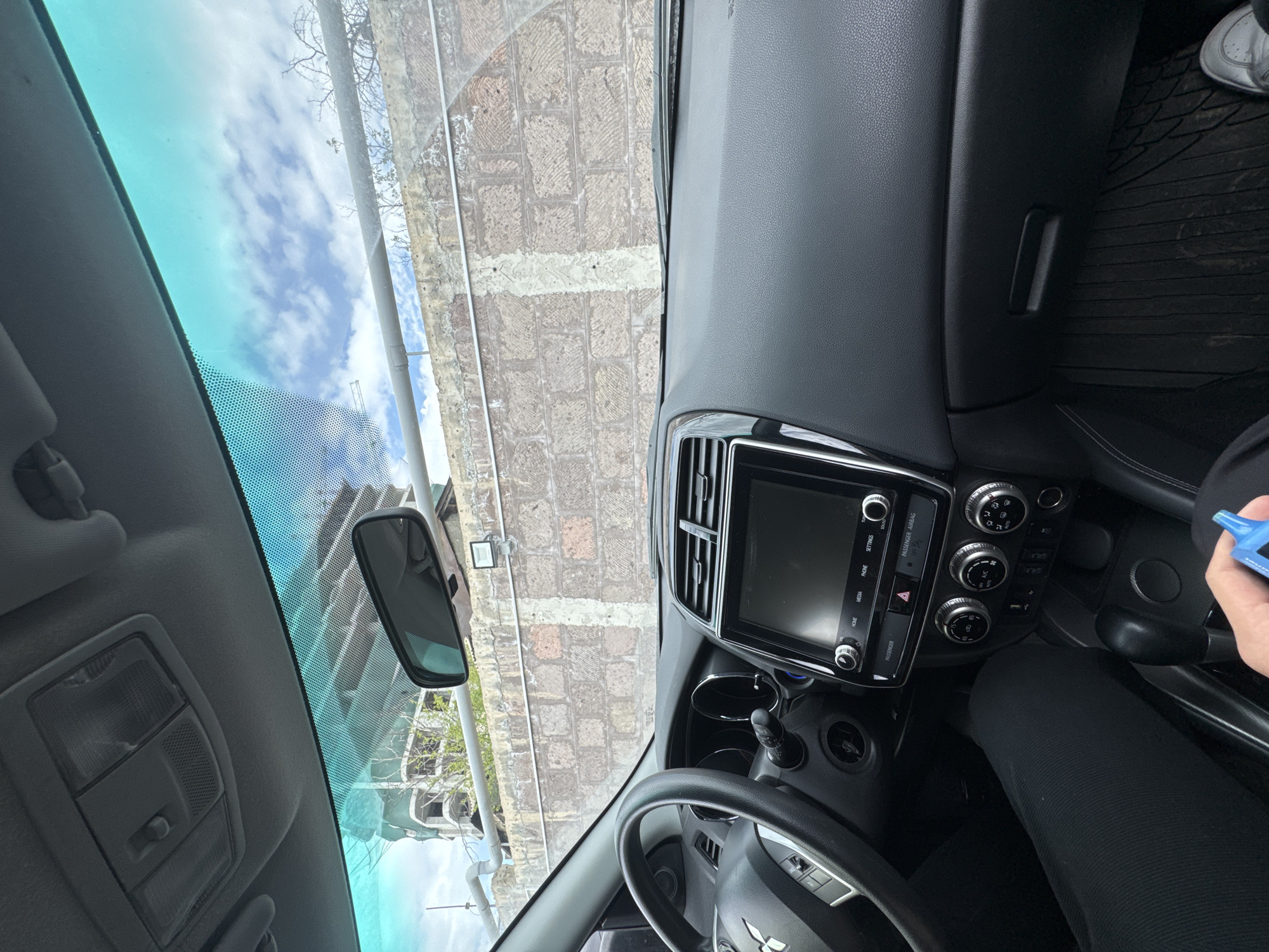 A first-person view from inside a car, likely a Mitsubishi based on the steering wheel emblem. The car is parked with a view of a brick building, a street, and sky with clouds visible through the windshield. The car's dashboard, steering wheel, center console with a touchscreen, and air conditioning controls are visible. A hand holding a small blue object is also present. The location is Yerevan, Armenia, based on the buildings visible in the windshield.