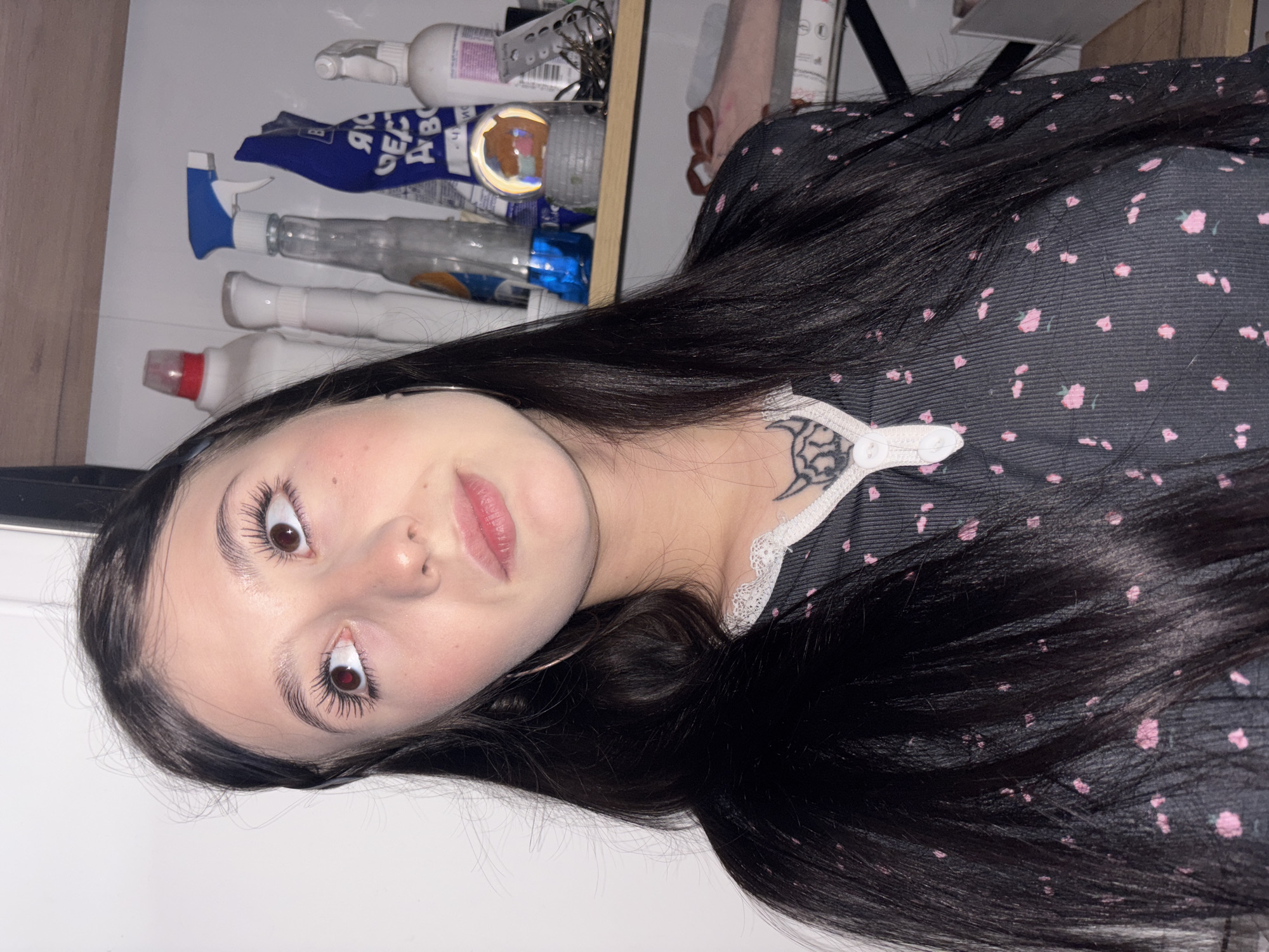 A young woman with long, dark, straight hair is shown in a close-up portrait, positioned horizontally in the frame. She looks upwards directly at the camera with wide, dark brown eyes enhanced by long, dark eyelashes. Her expression is attentive, with a soft pout on her pink lips and a light blush visible on her cheeks.
She is wearing a dark top adorned with a subtle pink floral pattern. A delicate white lace trim is visible around her neckline, and beneath it, an intricate dark tattoo, possibly a spiderweb design, is seen on her upper chest/shoulder. A thin metallic hoop earring is visible in her left ear. Her dark hair cascades around her head and over her shoulder, contrasting with her fair skin.
The background reveals a cluttered shelf filled with various household items, including several bottles and spray bottles. Some of these containers display partial Cyrillic text, such as "ЯР", "ДЕЗ", and "А В", suggesting a Russian-speaking context, consistent with the specified location of Фоки, Russia. Among the items is a clear spherical object with a visible reflection. The lighting appears to be artificial and bright, illuminating the indoor scene.