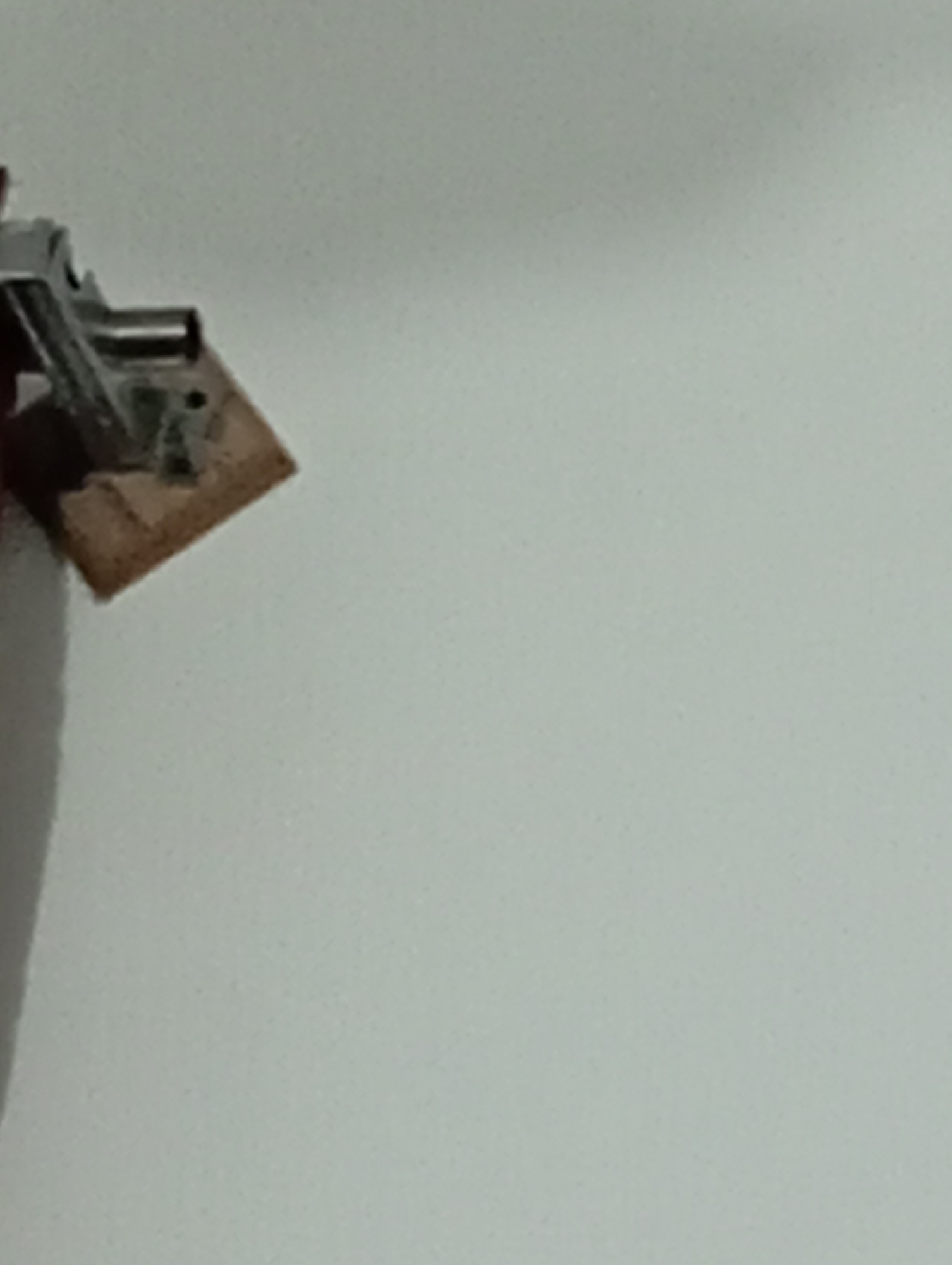 A close-up, overhead shot of a small, angled piece of wood with a metal clamp attached to its top surface. The wood is light brown and appears to be somewhat textured. The clamp is silver and has a hinged arm. The background is a plain, light gray surface, possibly a wall or a table. No location in Lassan, Pakistan is identifiable from this image.
