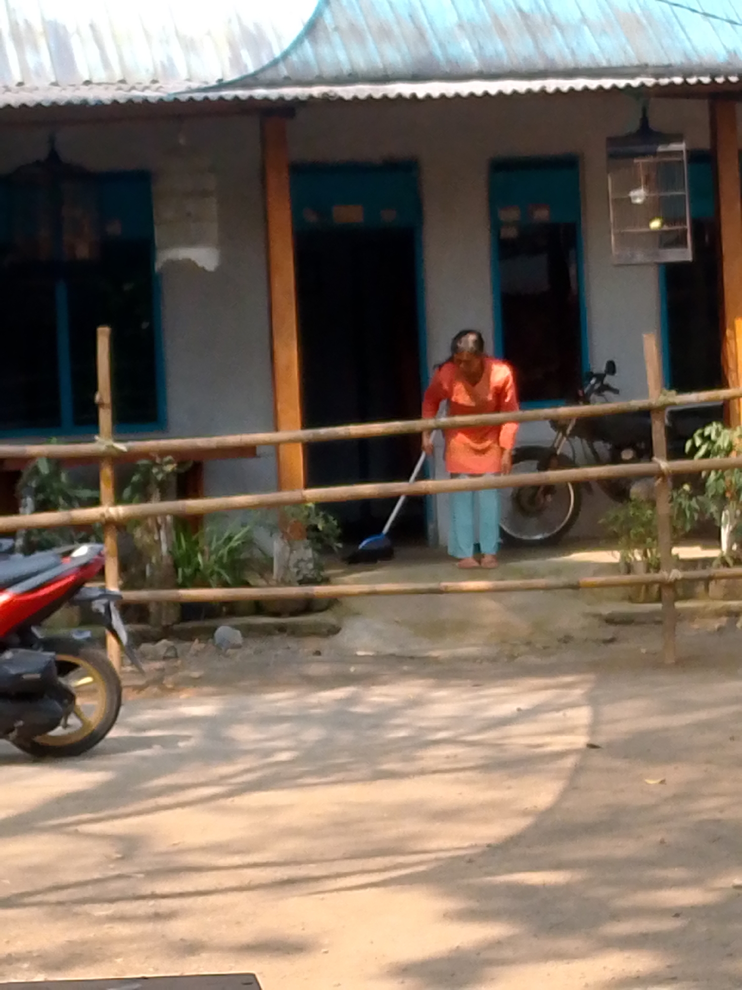 The image depicts a scene in front of a modest building, possibly a residence, in Pronojiwo, Indonesia. A woman, wearing an orange-red top and light blue pants, is sweeping the ground with a broom. She appears to be of middle age. To her right, a black motorcycle is parked. Behind the woman and the motorcycle is the building, characterized by a corrugated metal roof and light-colored walls. Two prominent windows with blue frames are visible, as is a birdcage suspended from the overhang of the roof. A bamboo fence runs across the foreground of the image, separating the building area from a dirt pathway. A red and black motorcycle is partially visible on the left side of the frame. The environment appears to be outdoors, with shadows suggesting it is daytime.