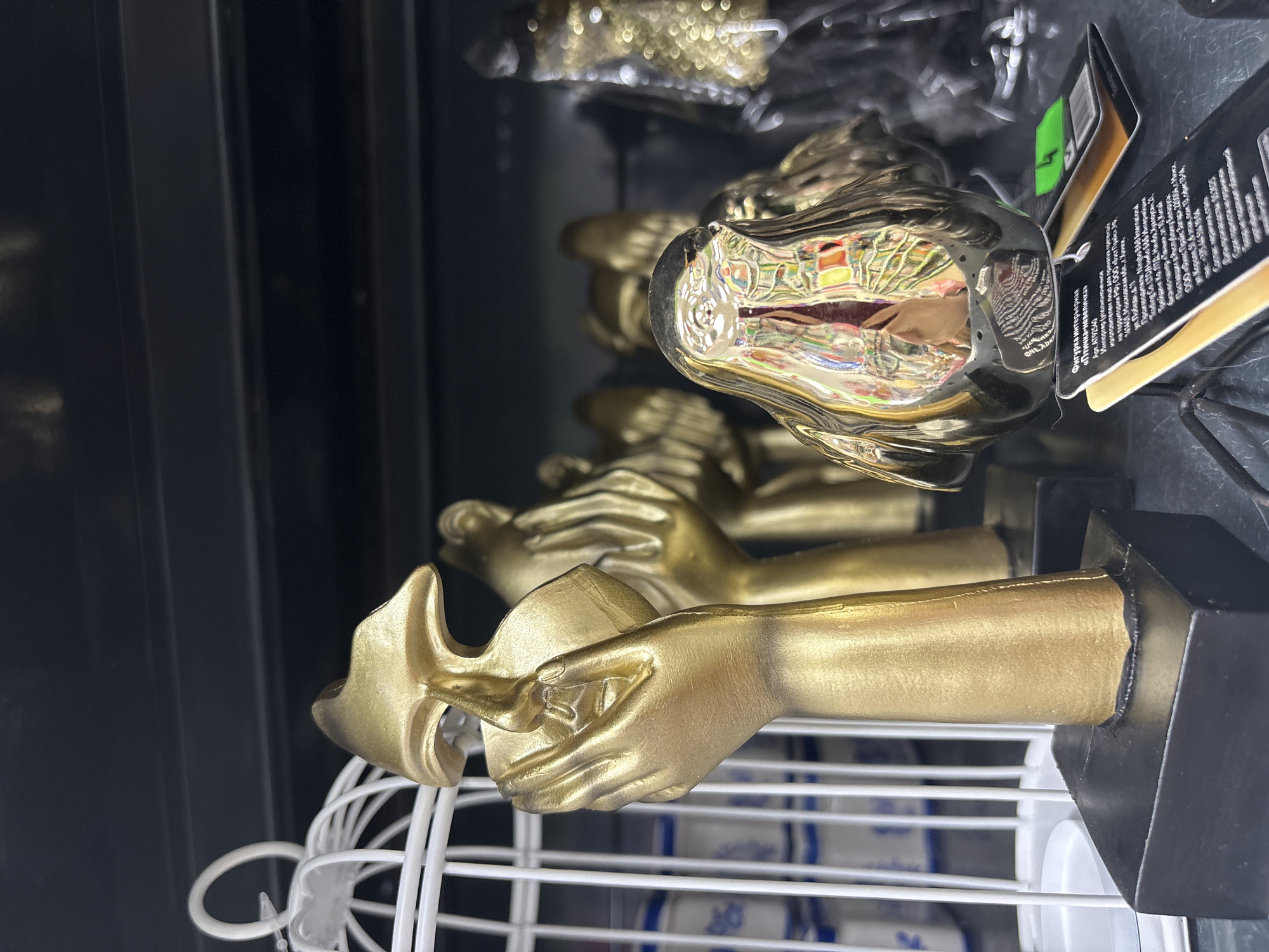 The image, oriented vertically, displays an array of golden decorative items, likely within a retail store in Chaikovsky, Russia. The scene captures a section of a product display, illuminated by artificial indoor lighting.
The main subjects are several identical golden figurines, each depicting a stylized human hand holding a sleek, mask-like or bird-shaped object. These sculptures are arranged on a white wire display rack in the foreground. The hands are detailed, showing visible fingers and knuckles, and the entire figurine has a consistent golden, somewhat matte finish.
Towards the upper right, another type of golden object is visible, distinct from the hand figurines. This item possesses a highly reflective, chrome-like surface, which distorts and mirrors various colorful lights and patterns from the surrounding environment. Attached to this particular item is a black and white product tag.
The background is largely dark and out of focus, indicating further shelving or the general interior of the store. Below the wire rack, glimpses of other products with blue and white packaging are faintly discernible.
The product tag provides significant textual information. It identifies the item as an "Interior figurine 'Little Bird'" (Фигурка интерьерная «Птичка-невеличка»). The tag lists the importer in the Russian Federation as ООО «Бэст Прайс» (LLC "Best Price") located in Khimki, Moscow region, and the manufacturer as Ningbo B&B Trading Co. LTD from Ningbo, China. It also mentions a supplier in the Republic of Belarus, ООО «Фикс Прайс Загадка» (LLC "Fix Price Zagadka") in Minsk. The composition is stated as polyresin and steel, with an unlimited shelf life and a production date of 15.11.2005. This information suggests the image was captured in a store like Fix Price, a common discount retailer in Russia, aligning with the Chaikovsky location.