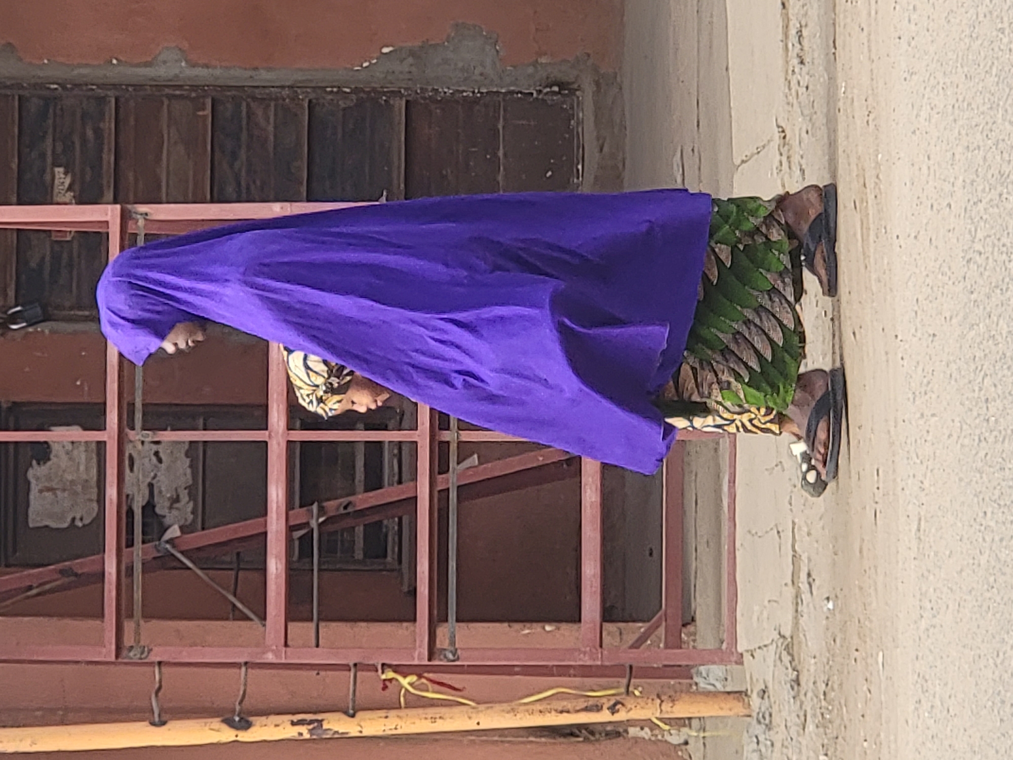 A woman stands in front of a building in Maiburiji, Nigeria. She is wearing a green patterned dress and a large purple headscarf or veil that covers much of her upper body. She also wears sandals. The building behind her has a reddish-brown window frame with metal bars over the windows. The building appears to be in a state of disrepair with peeling paint. There is a yellow metal pipe or bar running horizontally across the bottom of the frame. The ground is a light beige color.