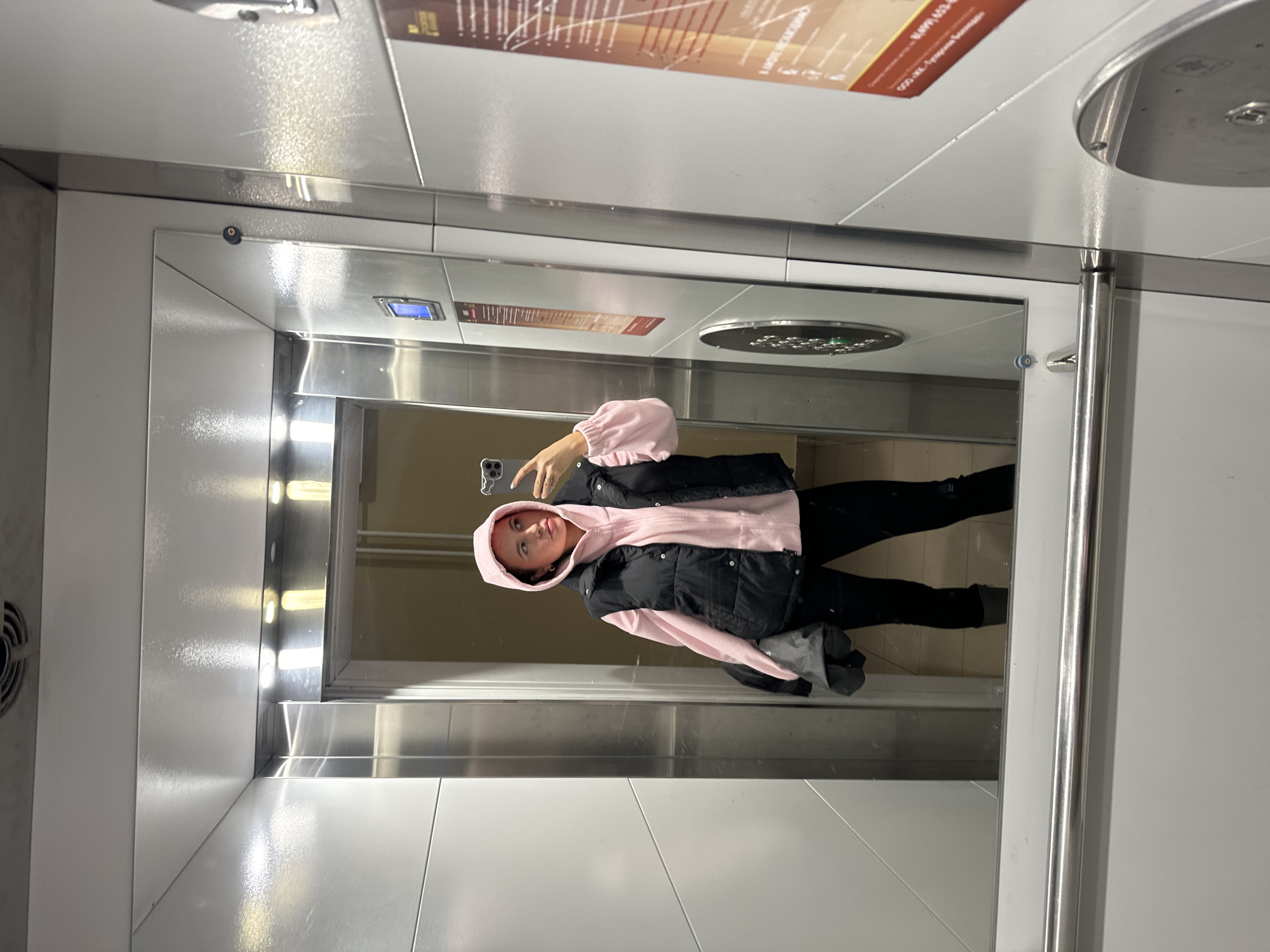 A female individual is visible in a self-portrait taken within an elevator cab. The location is an elevator in the Basmanny District of Moscow, Russia, as indicated by a poster on the right wall for "Management Company Basmannaya" (Управляющая компания Басманная) with a Moscow phone number.
The female, with light hair and fair skin, is wearing a light pink hooded sweatshirt with the hood up, a black sleeveless puffer vest over it, and black trousers. She holds a light-colored smartphone with a grey case in her raised right hand, aiming it towards the mirror to capture the image. A dark-colored garment is held in her left hand.
The elevator cab features a large reflective mirror on the left wall, capturing the full reflection of the individual. The interior walls are primarily composed of light grey or off-white panels, accented with polished stainless steel trim. The ceiling is light-colored with multiple recessed lights and a circular ventilation fan. A silver-colored horizontal handrail is mounted below the mirror. On the right wall, a control panel includes a circular array of buttons (some illuminated green), a rectangular blue-lit digital display, and another circular fixture below it. A poster with Russian text and graphics, related to a communal services management company, is affixed to the right wall. The floor visible inside the elevator appears dark, while the reflection from outside the elevator doors suggests a lighter-colored floor in the building's corridor.