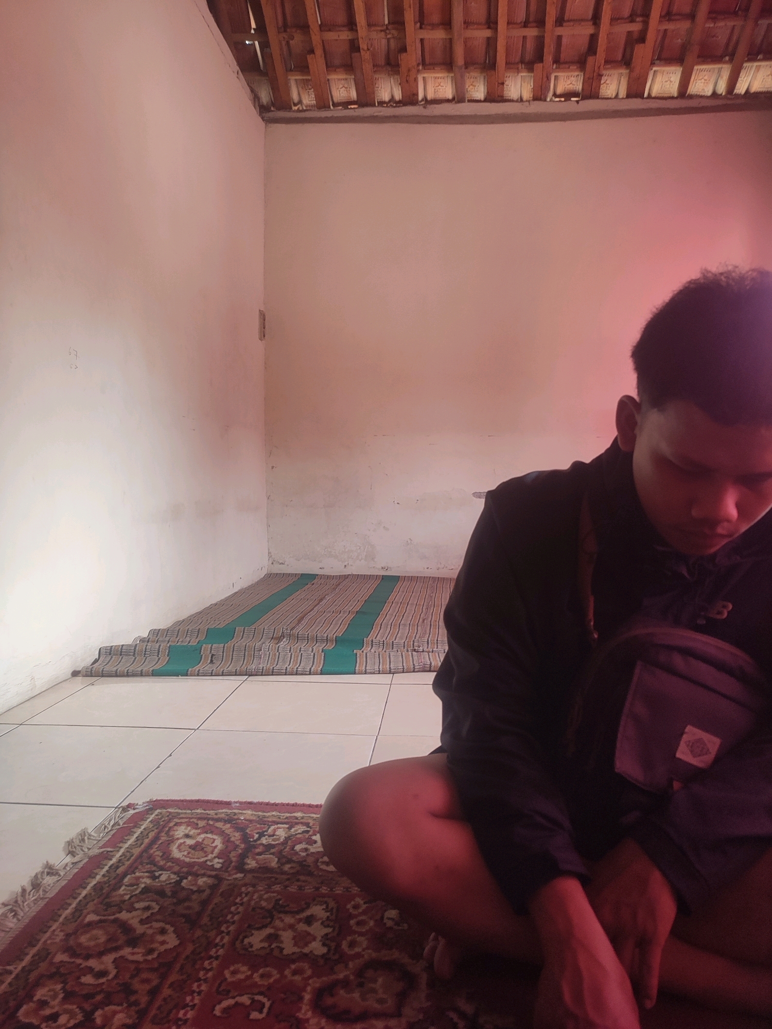 A young man is sitting cross-legged on a red patterned rug in a room. He is wearing a black jacket and a grey bag is slung across his chest. The room has white walls, a tiled floor, and a wooden ceiling. A striped mat with green and brown stripes is laid out on the floor. The overall lighting in the room is dim, with a pinkish hue. The image was taken in Karangtuang, Indonesia.