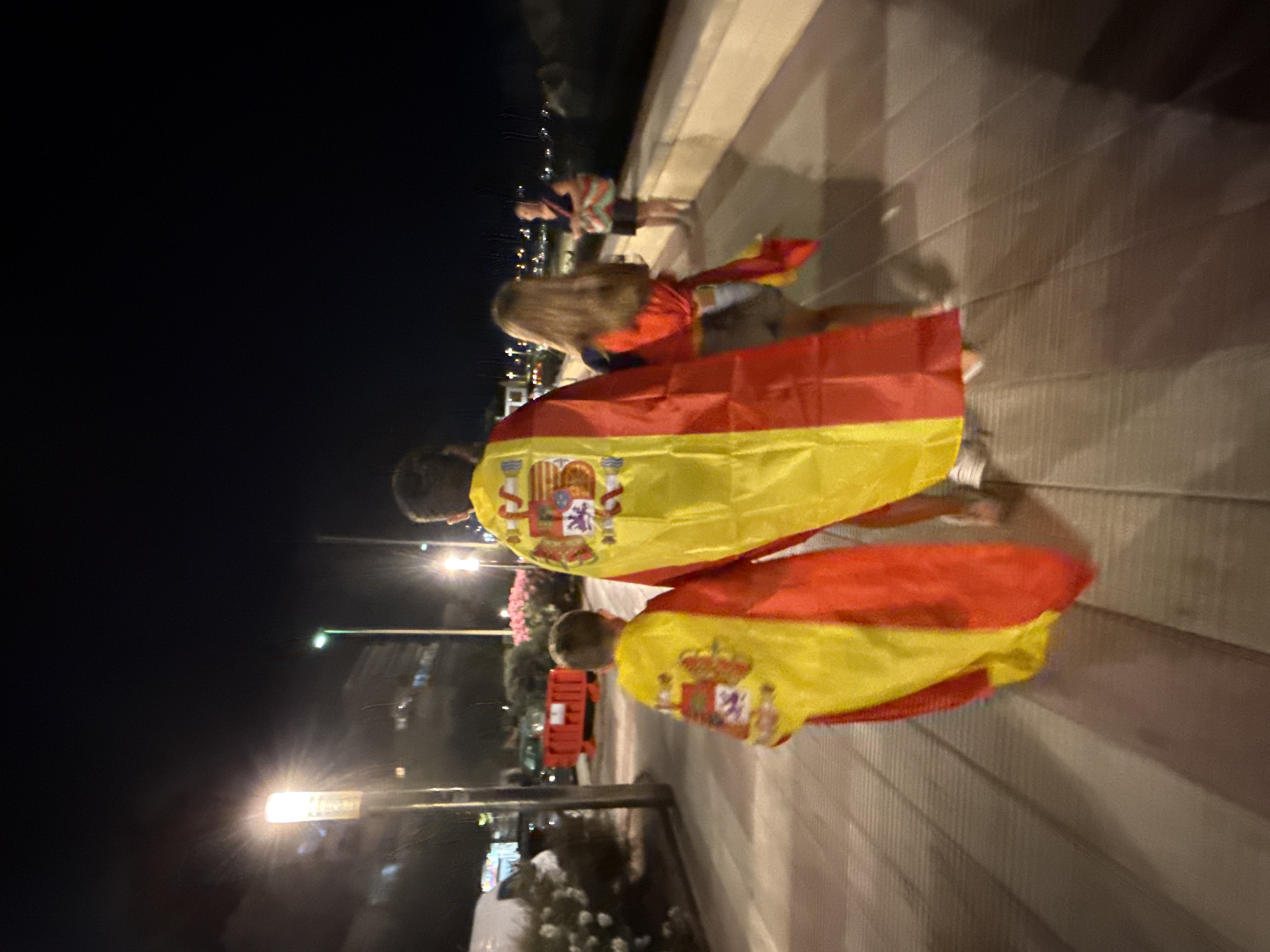 Two individuals, one adult and one child, draped in Spanish flags, walk away from the camera on a paved promenade at night. The child, in the foreground, is wearing a Spanish flag like a cape. Behind them, an adult also has a Spanish flag draped over their shoulders. To the right, a woman with blonde hair, wearing shorts and a top, walks towards them, also partially draped in a Spanish flag. Further in the background, another woman stands near a low wall. The environment is a city promenade with streetlights and some vegetation visible. The scene is set in Blanes, Spain.