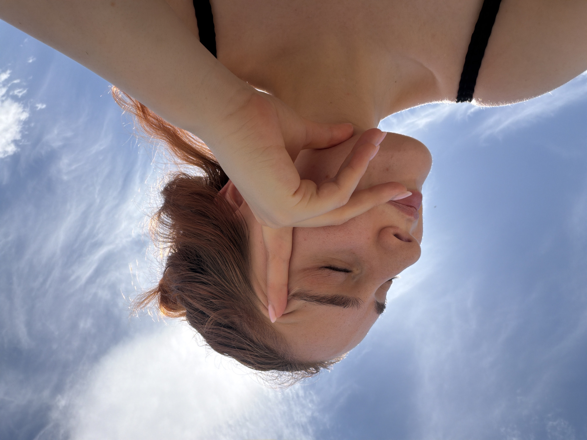 The image is a low-angle, close-up shot of a young woman's face and upper body against a bright blue sky with wispy clouds. The woman has her eyes closed, and her right hand is resting on her forehead, with her thumb touching her temple and her fingers extended upwards. Her hair is a reddish-brown color and is styled loosely, with some strands catching the light. She is wearing a black top. The lighting suggests it is daytime, and the sun is likely behind and to the left of the frame, creating a slight lens flare effect. There are no identifiable landmarks or clues within the image that would indicate a specific location in Strasbourg, France. The scene is focused on the woman's expression and the natural light.