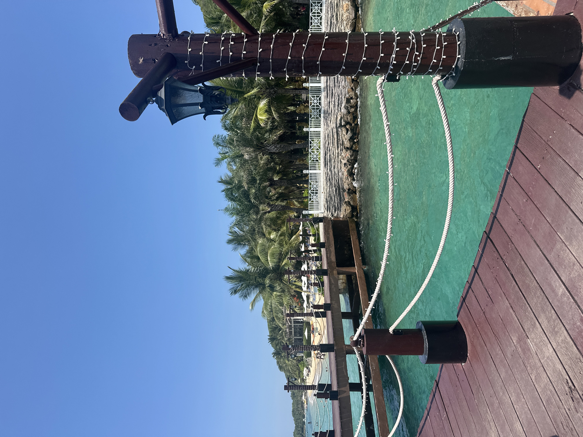 This image depicts a coastal scene on a sunny day in Nha Trang Ward, Vietnam.
In the foreground, a dark reddish-brown wooden plank pier extends from the lower left towards the mid-ground. On the right side of the pier, a prominent dark brown wooden post, wrapped with a spiral of small white LED string lights, stands at the edge. A black, ornate, multi-faceted glass lantern is attached to a horizontal dark brown wooden beam that extends left from the top of this post. Two strands of thick white rope, intermittently wrapped with string lights, extend horizontally from the post, forming a railing along the pier's edge. The post is anchored in a black cylindrical base.
The mid-ground features a body of clear, vibrant turquoise water directly beneath and to the left of the pier. Further into the scene, another section of the wooden pier extends towards a sandy beach, also equipped with similar brown posts and rope railings.
In the background, a sandy beach is visible, populated with numerous white sun loungers and umbrellas. Several individuals are present on the beach and in the shallow water, some appearing to be swimming or wading. A dense line of tall, green palm trees borders the beach and extends up a verdant, tree-covered hill on the right. A white decorative fence or balustrade runs along a paved path on the shoreline, separating the water from a cluster of palm trees and tropical foliage. Portions of white-structured buildings, consistent with a resort, are discernible through the trees.
The sky is a clear, bright blue, occupying the upper half of the image, indicating fair weather conditions. The overall scene suggests a tranquil, tropical environment.