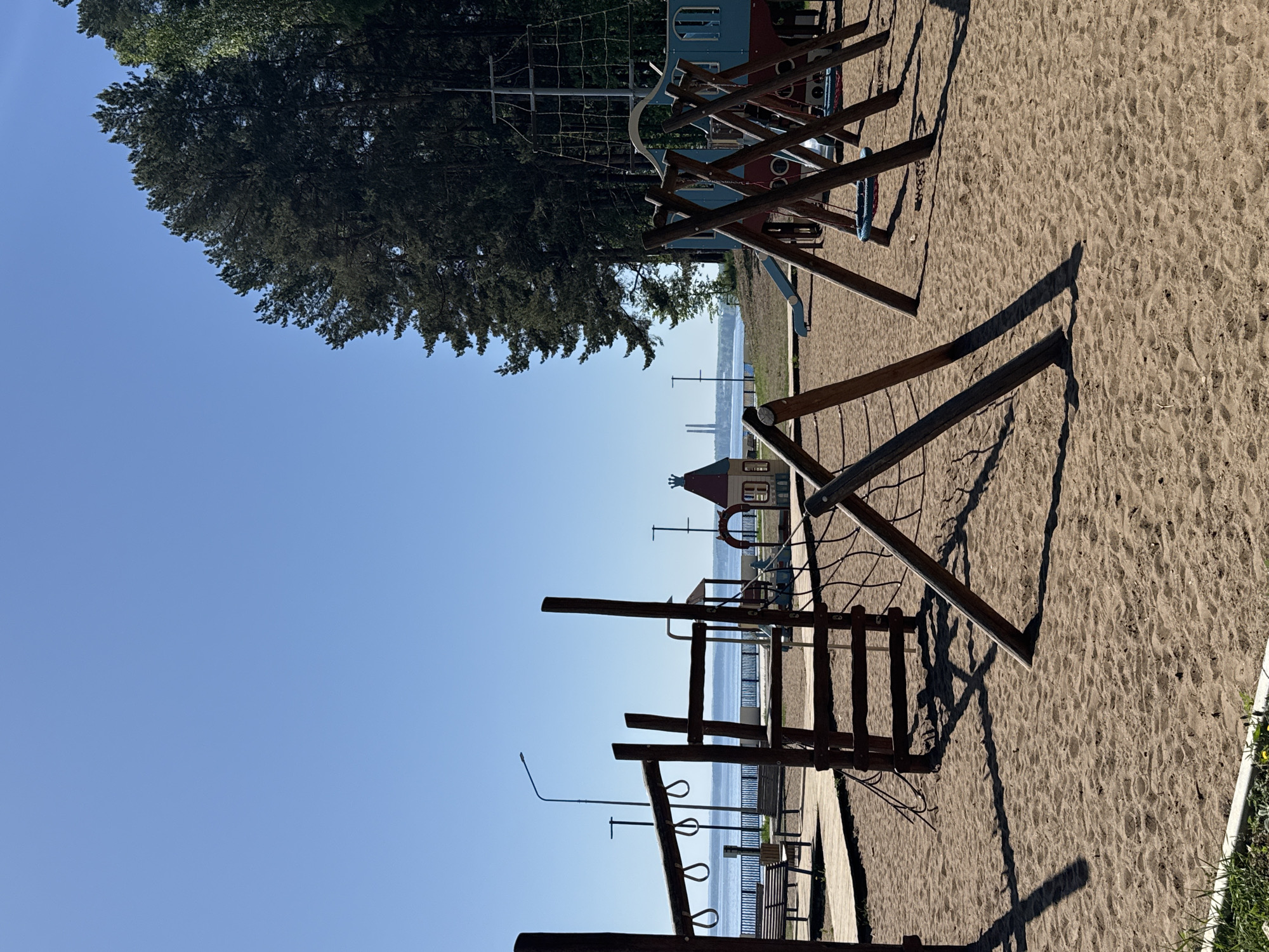 The image shows a playground located in the city of Chaikovsky, Russia. The playground has several wooden play structures in a sand-filled area. There are climbing structures, swings, and a playhouse. The Volga River can be seen in the background, along with benches and streetlights along the riverbank. The sky is clear and blue. The photo seems to have been taken in the early morning.