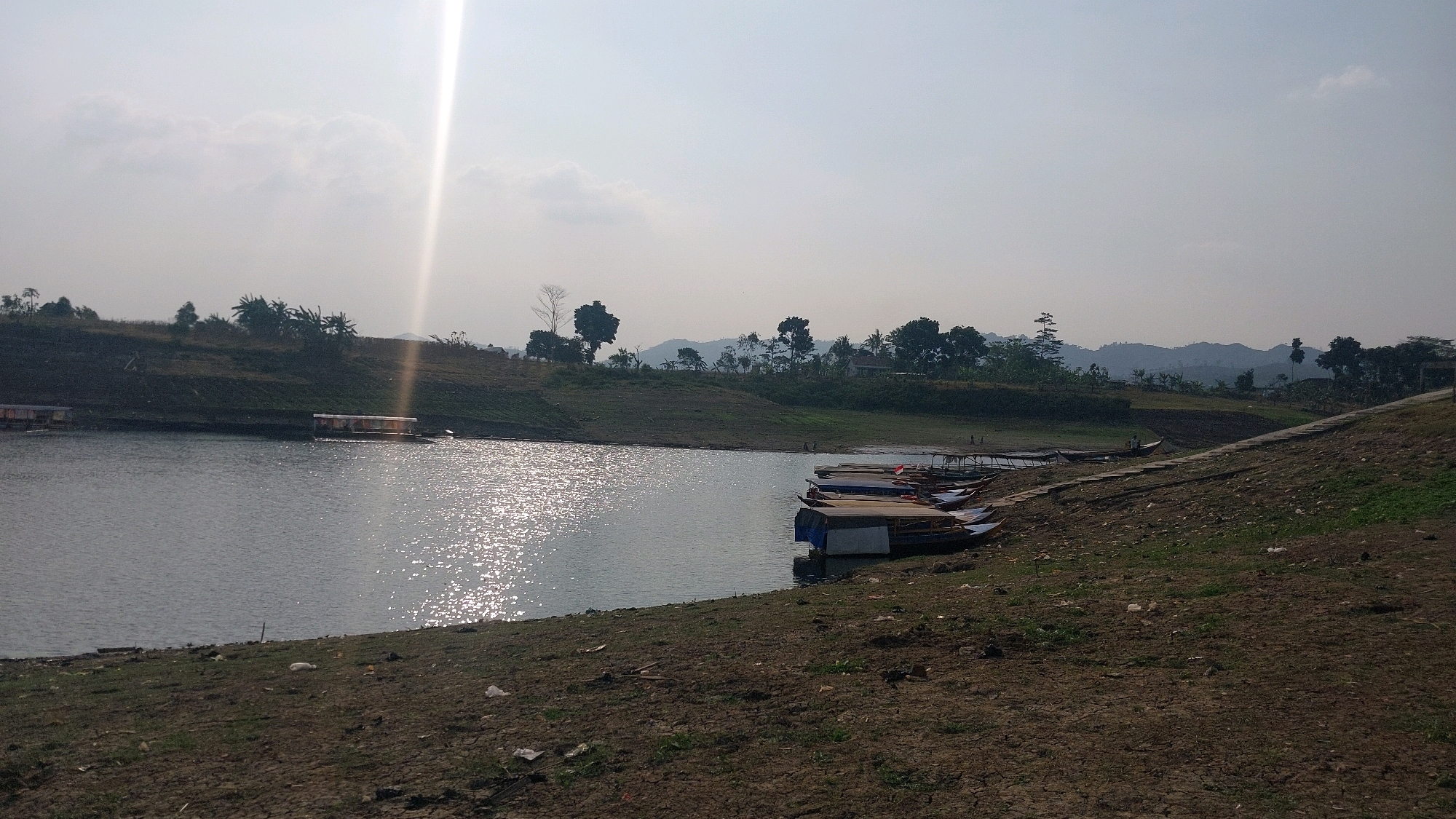 The image depicts a sun-drenched landscape at the location of Bojonghaleuang, Indonesia. A calm, vast body of water dominates the scene, reflecting the bright sunlight with numerous shimmering patches. The water's edge on the right side of the frame is a dry, cracked earth embankment sparsely covered with dry grass and scattered litter. A paved pathway runs along this embankment, leading towards the background where rolling green hills are visible. Several boats are moored along the left bank of the water, some appearing as simple rafts with covered seating areas, while others are sleeker, more traditional boats. In the distance, across the water, a lush green hillside rises, dotted with trees and vegetation. The sky is bright and hazy, with a strong sunbeam piercing through from the upper left, creating a lens flare effect. The overall impression is one of a serene, possibly rural, waterfront area.