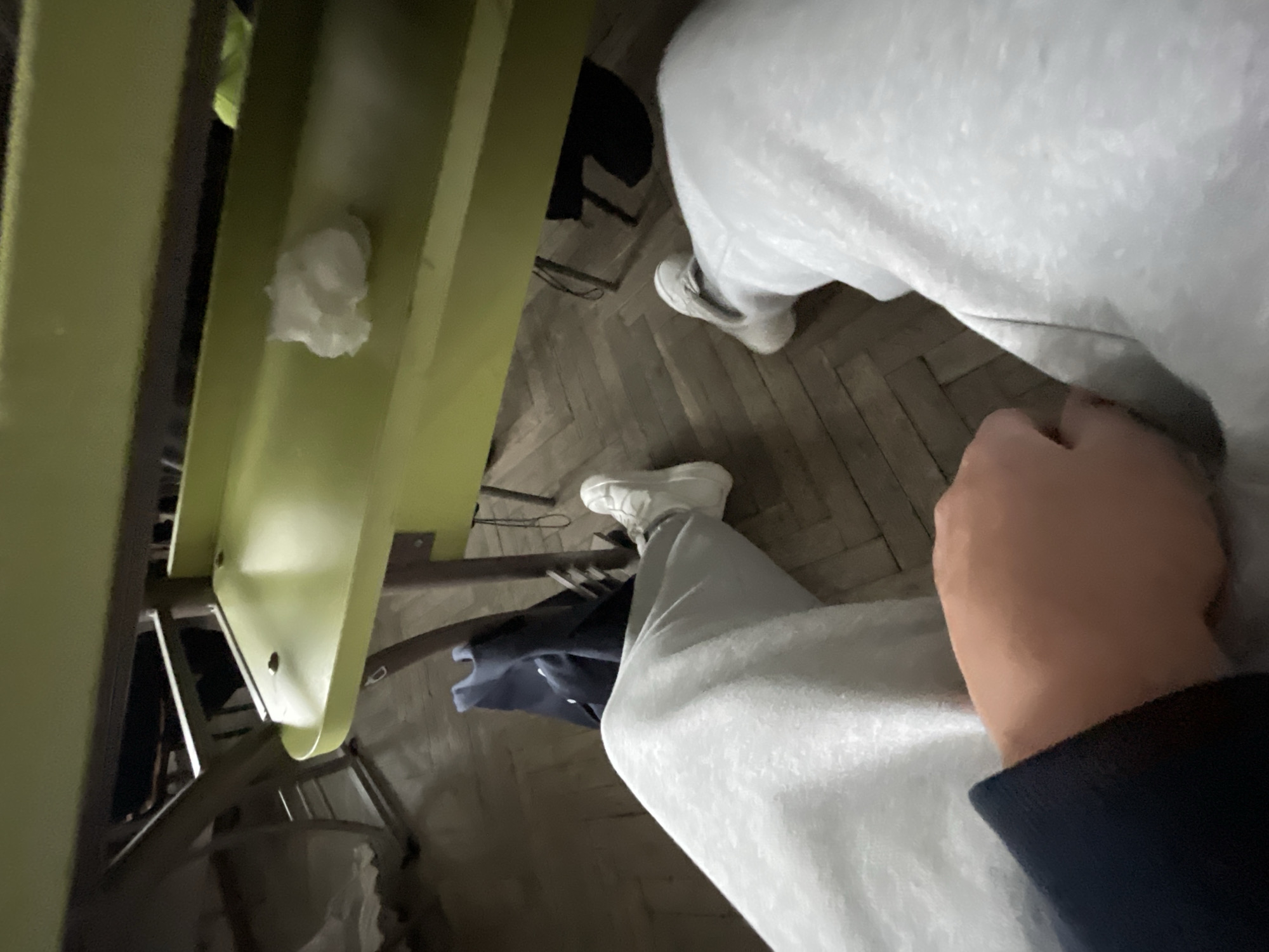 A first-person perspective image shows a person sitting in what appears to be an old-fashioned chair in a room with a herringbone-patterned floor. The person is wearing white sneakers and light grey sweatpants, and part of a dark blue sleeve and hand are visible in the lower right corner. The chair is painted light green and has a piece of crumpled white material attached to it. The location is unknown but hypothesized to be Zlatograd, Bulgaria.
