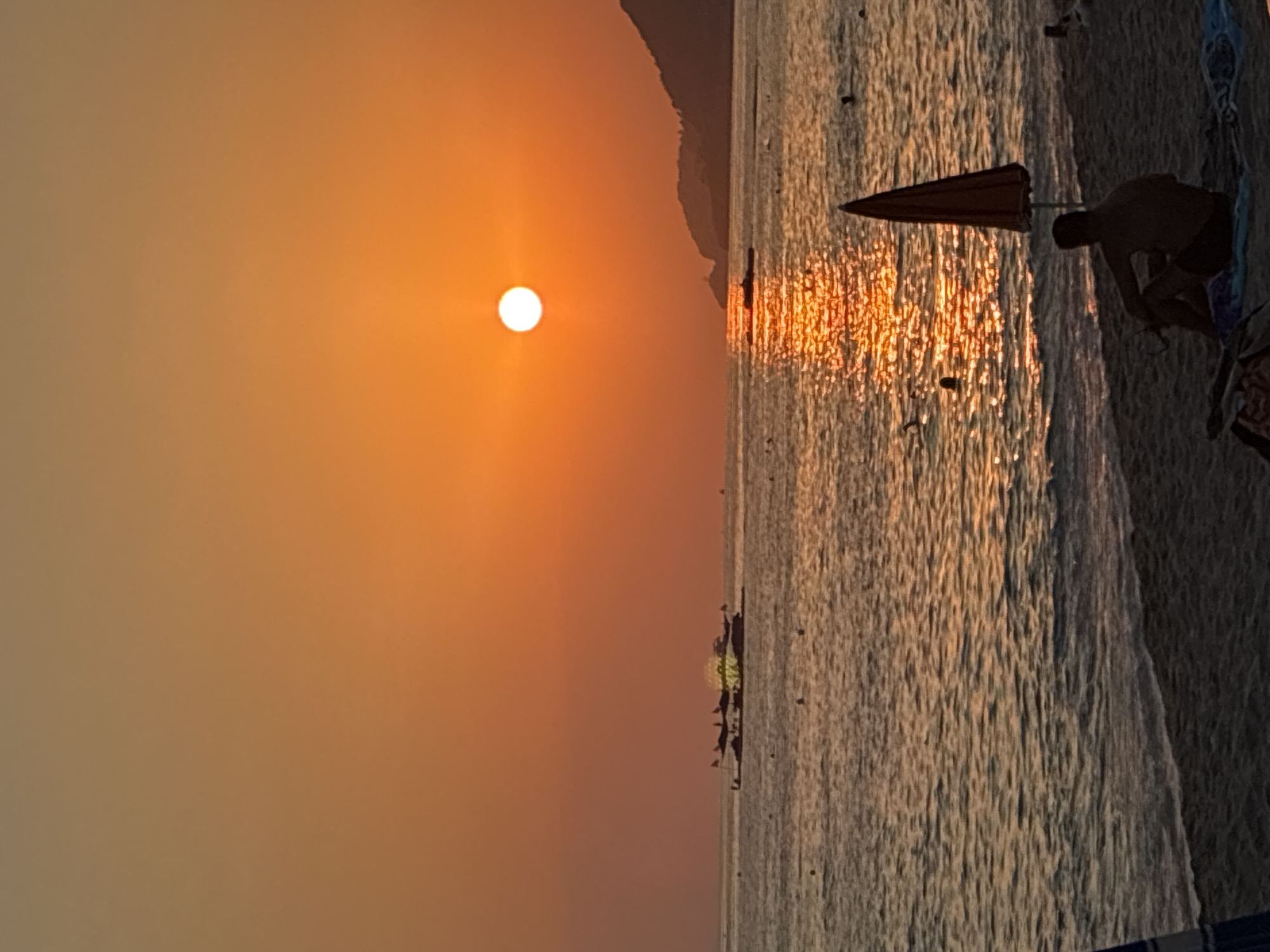 The image depicts a tranquil sunset scene at a beach in Alanya, Turkey. The sun, a bright white disc, is low on the horizon, casting a warm, orange glow across the sky and water. The sea is calm with gentle waves lapping at the shore. In the distance, several boats are visible, silhouetted against the vibrant sky. On the beach, a lone figure sits under an umbrella, gazing out at the sea. The overall atmosphere is one of peace and relaxation.