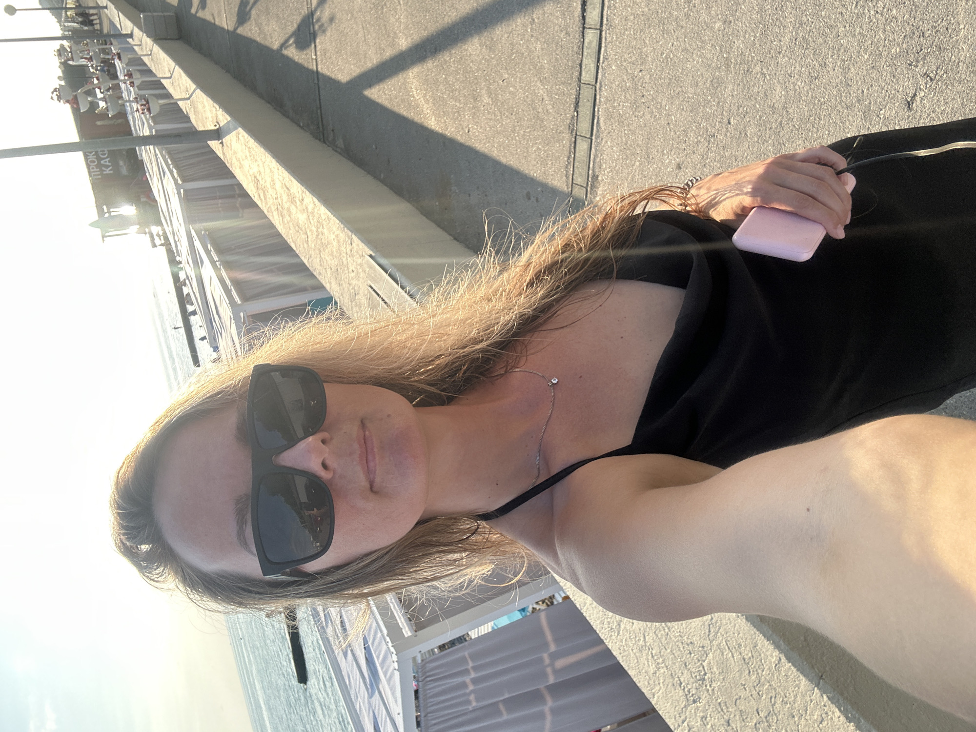 This is a selfie of a woman on a paved walkway, possibly a promenade, next to a body of water. The woman is wearing sunglasses and a black top, and she is holding a pink object, likely a phone or power bank, in her right hand. Her hair is long and light brown. The sun is low in the sky, casting long shadows and creating a warm, golden light, suggesting it is either early morning or late afternoon. In the background, there are several white structures that appear to be beach cabanas or kiosks, and beyond that, the ocean. There is some text visible on one of the structures in the background, appearing to be in Cyrillic. The overall mood is relaxed and summery.