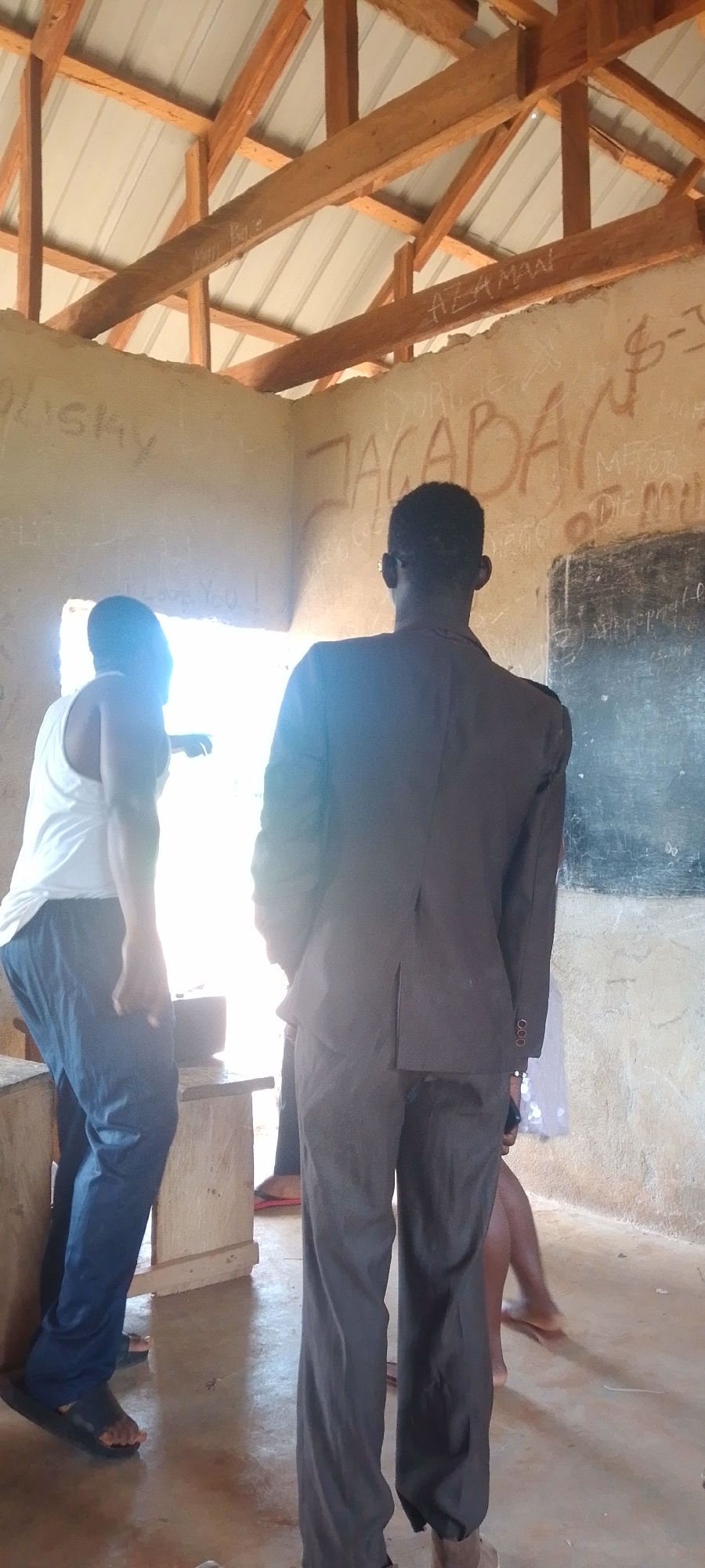 The image shows the interior of a room with rough, unplastered walls and a wooden beam ceiling. The walls are covered in graffiti, with some legible words like "AZAMAN" and "JACABADAN" visible. A blackboard is mounted on the far wall. There are at least three people visible. In the foreground, a man in a dark suit stands with his back to the camera, facing away from the viewer. To his left, another person, wearing a white tank top and blue pants, is seen from the side, pointing towards an opening that lets in bright sunlight, suggesting it might be daytime. A third person's legs are visible in the lower right of the frame, and a fourth person's legs are partially visible in the lower left. The overall impression is of a rustic or unfinished indoor space, possibly a classroom or community building. The bright light from the opening could indicate a sunny day. The graffiti suggests a place where people have gathered and left their mark.